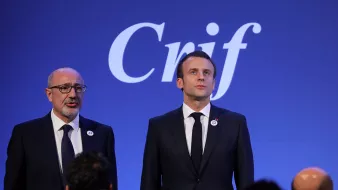 The image shows two men standing side by side in front of a blue backdrop with the word "Crif" displayed prominently. One man is dressed in a black suit and tie, appearing to be delivering a speech or presentation. The other man, also in a formal suit, stands next to him, possibly in a supportive or attentive role. The setting suggests a formal event, likely related to the organization represented by "Crif."