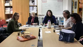 The image depicts a group of five women sitting around a table in a room filled with shelves and papers. They appear to be engaged in a discussion or work-related activity. The table has some items on it, including drinks and papers, suggesting a collaborative atmosphere. The women are looking at each other, indicating communication and interaction among them. The setting seems to be an office or a meeting room.