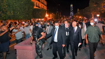 The image depicts a nighttime scene in a city, where a group of people is gathered in a street. In the foreground, a group of officials, including one dressed in a suit, is walking through a crowd, accompanied by a person in tactical gear, suggesting a security presence. The atmosphere appears charged, with some people in the crowd gesturing expressively. Trees and buildings with lights are visible in the background, contributing to the nighttime setting. Overall, the scene conveys a sense of political or social gathering, possibly in response to current events.