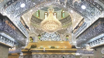 The image depicts the interior of a beautifully ornate religious site, characterized by intricate mirror work and elaborate decorations. The ceiling is adorned with shimmering tiles and a large chandelier, creating a luminous atmosphere. There is a golden structure at the center, likely a shrine or tomb, surrounded by a crowd of women wearing traditional black clothing, all appearing to be engaged in prayer or reflection. The walls and ceiling are richly decorated with calligraphy and other designs, enhancing the sacred ambiance of the space.