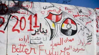 The image features a wall covered in graffiti and artwork. Prominent elements include the Egyptian flag, the date "25 January 2011," and references to the revolution. Phrases in Arabic and English express themes of hope, such as "Better Future" and "إرادة," meaning "will" or "determination." The background suggests a lively and politically charged atmosphere, symbolizing the spirit of change during the Egyptian revolution. Various colors like red, black, and white are used, enhancing the visual impact of the message.