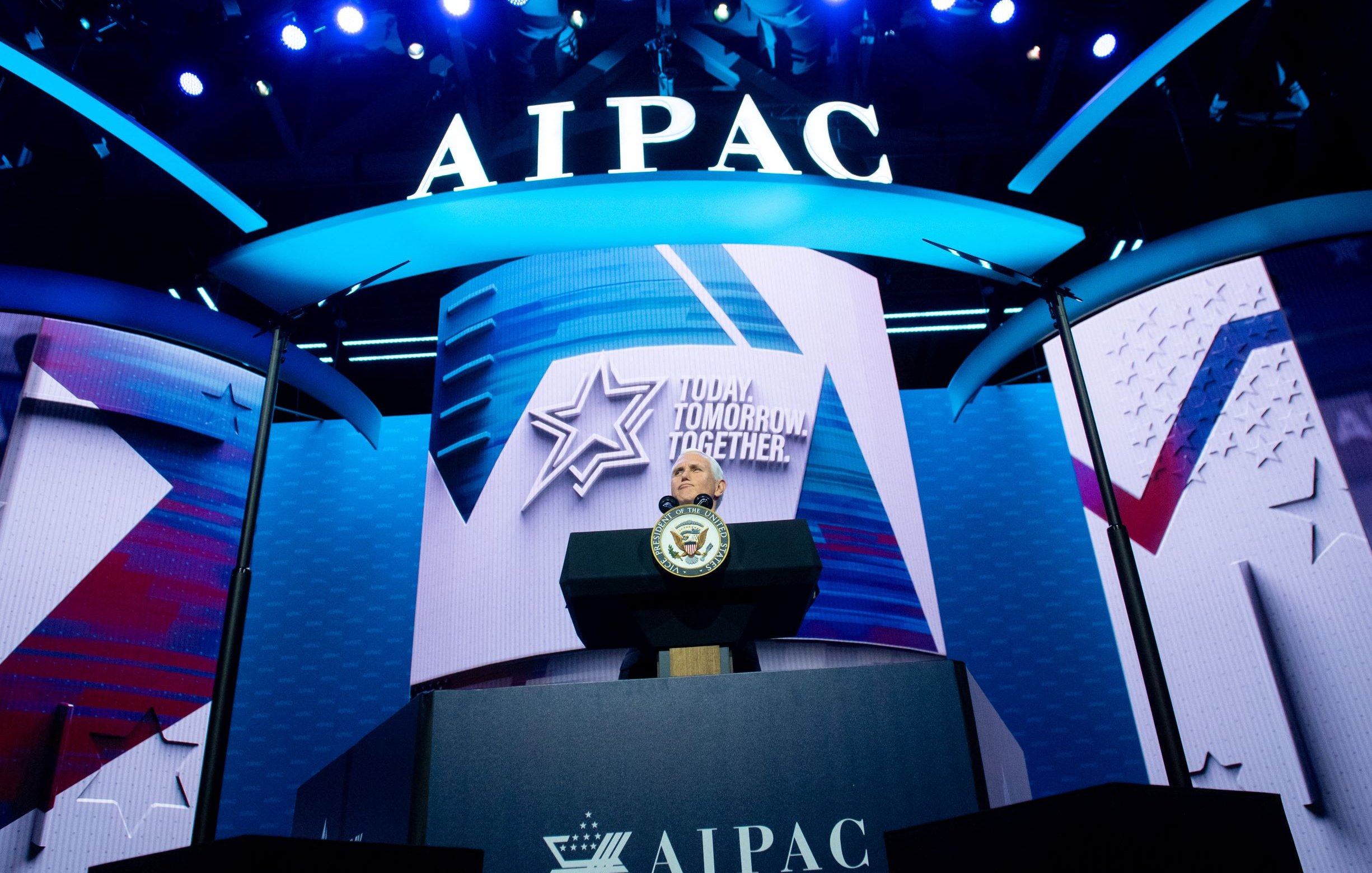 La imagen muestra un escenario iluminado con luces brillantes en un evento, con un gran fondo donde se puede ver el logo de AIPAC (Comité de Relaciones Públicas América-Israel). Hay un podio en el centro, probablemente destinado a un orador, con un sello oficial, indicando que se trata de un evento importante. La decoración incluye elementos gráficos como estrellas y símbolos que refuerzan un mensaje de unidad y futuro, destacado en el fondo.