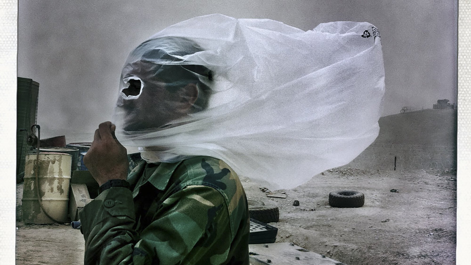 La imagen muestra a un hombre vestido con un uniforme de camuflaje, protegiéndose de una tormenta de arena. Su rostro está cubierto con una tela transparente que parece ser una especie de máscara o protección. El entorno se ve polvoriento y desolado, con algunos objetos y neumáticos dispersos en el área. La escena transmite una sensación de dificultad y adversidad debido a las condiciones climáticas.