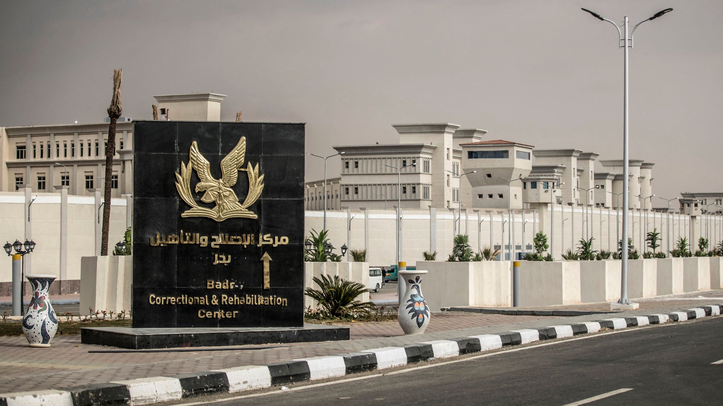La imagen muestra un letrero grande y elegante que indica la entrada del "Centro de Corrección y Rehabilitación de Badr". El letrero tiene un diseño prominente con un símbolo de un águila en la parte superior. Al fondo se puede ver una construcción moderna y amplia, posiblemente el centro mismo, rodeada de muros altos y bien cuidados. La carretera está pavimentada y hay algunas decoraciones de cerámica a los lados. El cielo parece nublado, dando un ambiente de calma.