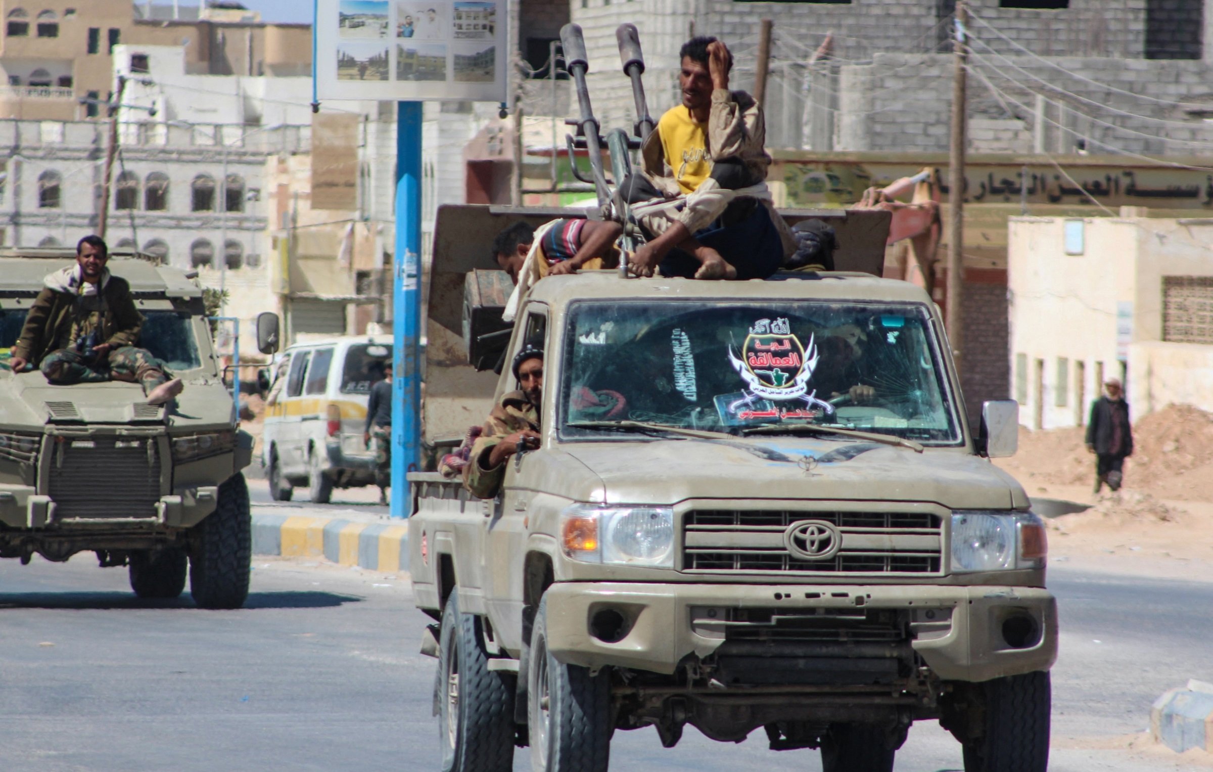 La imagen muestra un convoy de vehículos militares, principalmente camiones, en un ambiente urbano. En uno de los camiones, varias personas están sentadas y de pie en la parte trasera. Algunos están armados y parecen estar en posición de vigilancia. Los vehículos tienen un color beige o khaki, típico de vehículos militares. En el fondo, se pueden ver edificios y una carretera, lo que indica que están en una zona urbana. La escena transmite una sensación de actividad militar en la calle.