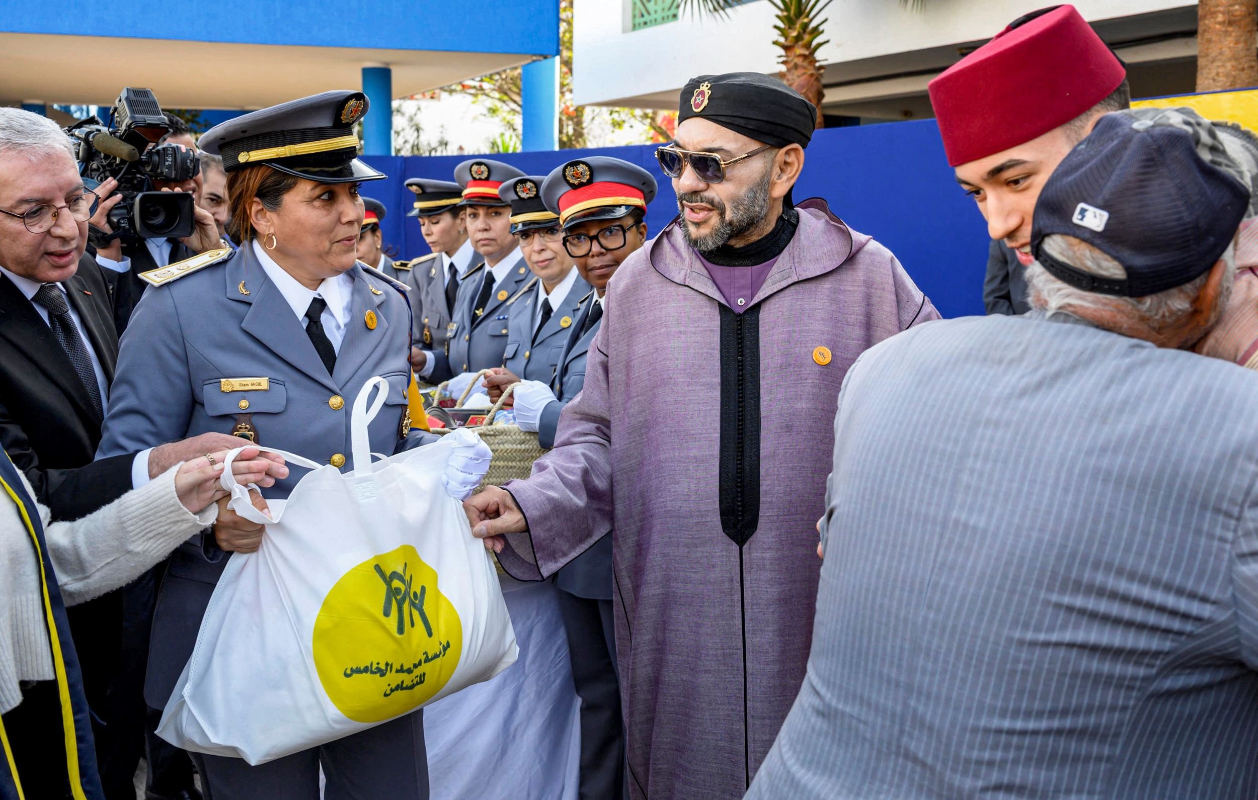 La imagen muestra a un grupo de personas en un evento ceremonial al aire libre. En el centro, hay un hombre con gafas y una túnica tradicional que parece estar recibiendo una bolsa de una mujer uniformada. A su alrededor hay varios otros individuos, algunos en uniformes y otros con gorros tradicionales. Todos parecen estar en una atmósfera de celebración o entrega de ayuda, y el ambiente está adornado con plantas y un edificio de fondo.