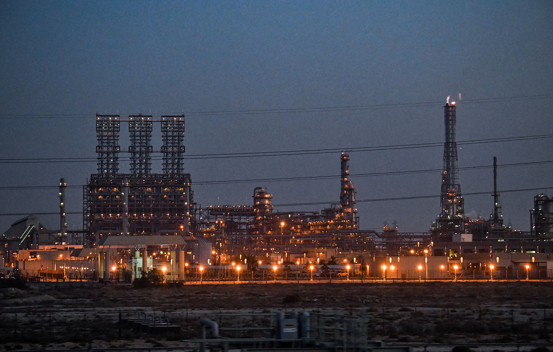 La imagen muestra una instalación industrial, probablemente una refinería o planta petroquímica, iluminada al anochecer. Se pueden ver numerosas estructuras metálicas altas, chimeneas y torres, con luces brillantes que resaltan en el entorno oscuro. Hay líneas eléctricas que cruzan la imagen y el paisaje es árido, lo que sugiere que la planta está ubicada en una zona desértica o poco vegetada. La atmósfera transmite una sensación de actividad industrial, con un enfoque en la tecnología y la producción.