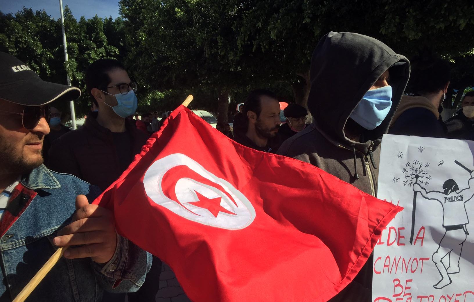 En la imagen se puede ver a un grupo de personas participando en una manifestación. Uno de los manifestantes sostiene una bandera roja con una estrella y una media luna blancas, símbolo nacional de Túnez. Otro individuo lleva un cartel que dice "Una idea no puede ser destruida", acompañado de una ilustración. La mayoría de los participantes utilizan mascarillas, y el ambiente parece ser de protesta pacífica. El fondo muestra árboles y un entorno urbano.