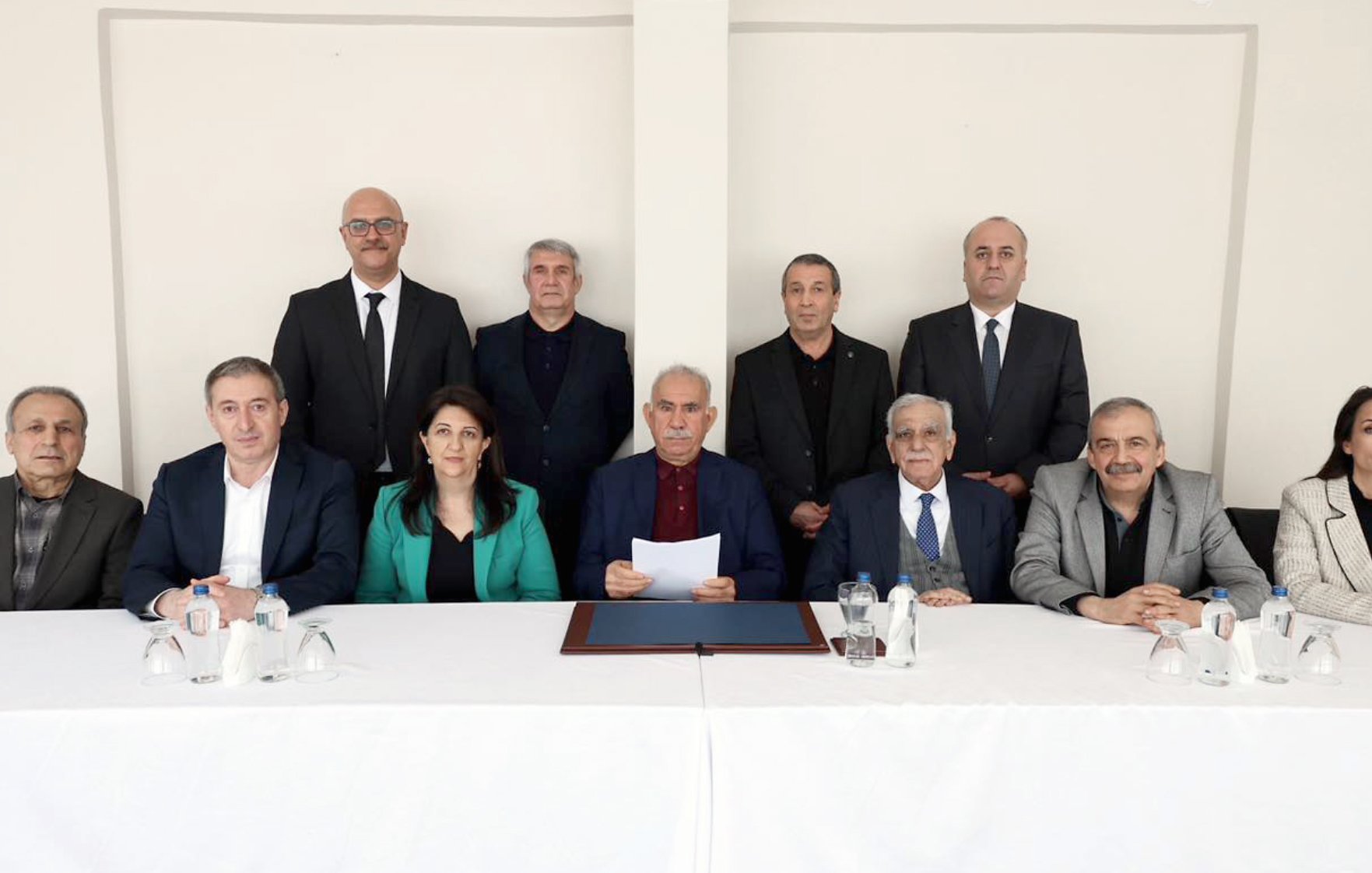 The image shows a group of twelve people seated around a table with a white cloth. They are dressed formally and appear to be engaged in a serious discussion or meeting.