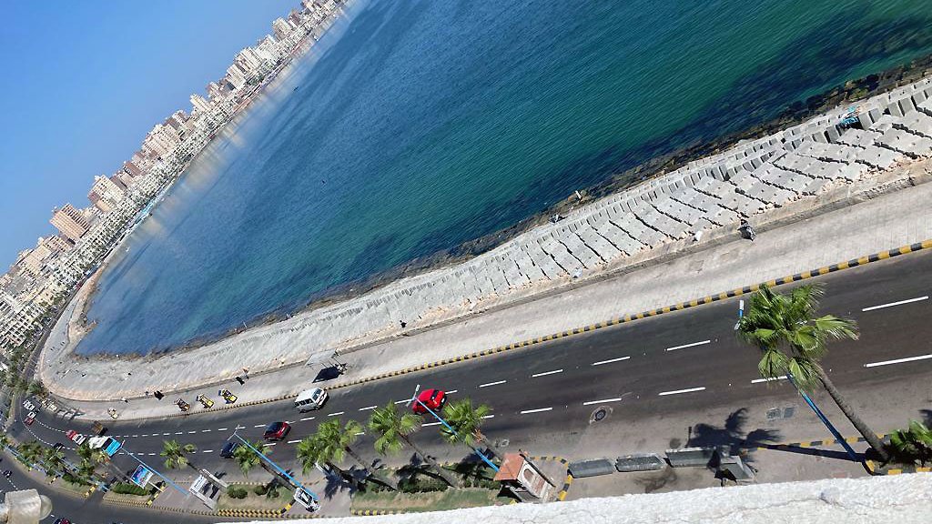 La imagen muestra una vista aérea de una costa bien iluminada. Se puede ver una carretera que corre paralela a la playa, con varios vehículos transitando. A lo largo de la playa hay palmeras, y el agua del mar es de un color azul claro. Al fondo, se pueden observar edificios que forman parte de un área urbana, y el cielo está despejado, lo que sugiere un día soleado.