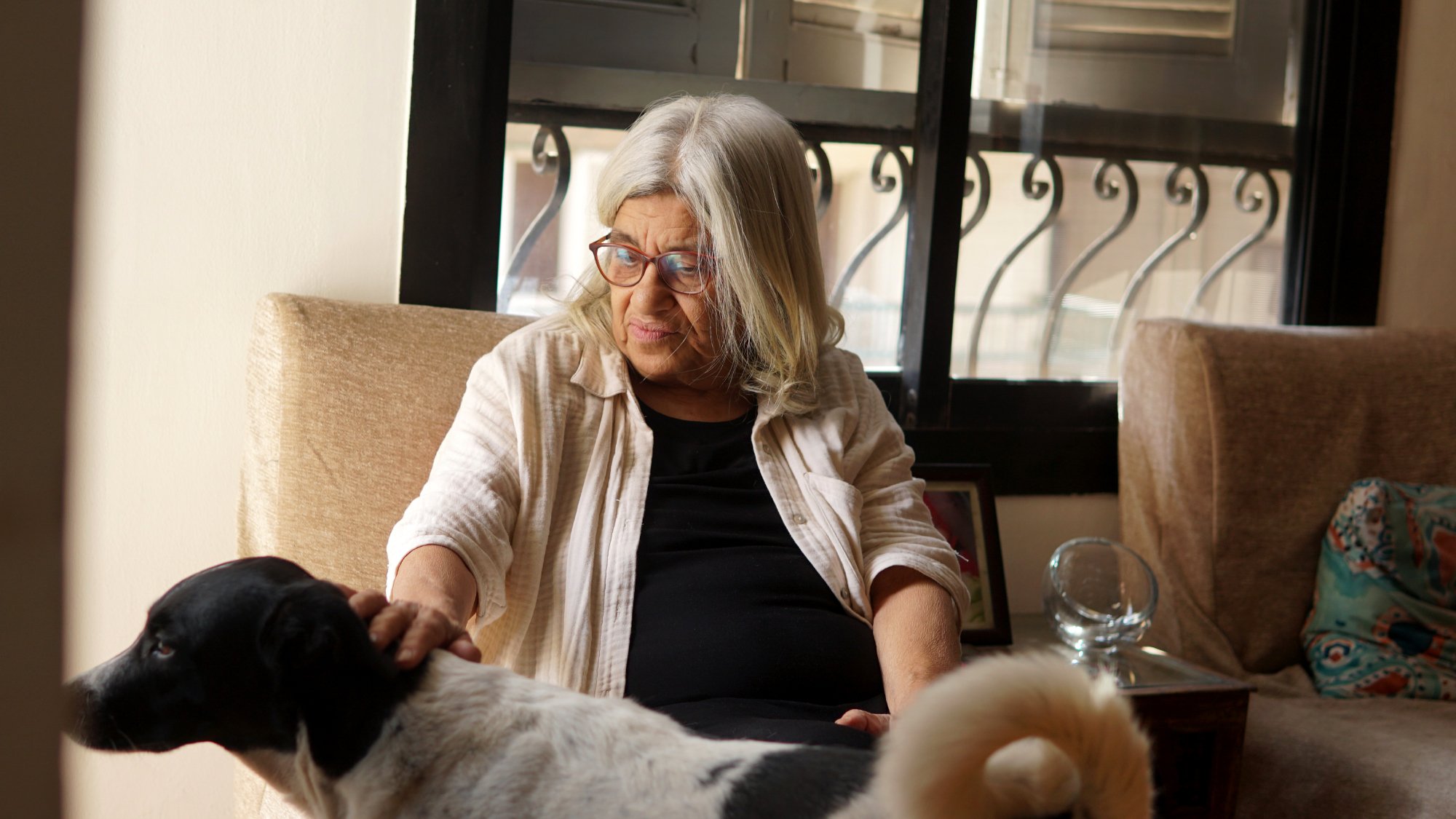 La imagen muestra a una mujer mayor sentada en un sofá, con cabello largo y canoso. Ella lleva gafas y viste una camiseta negra con una chaqueta clara. Está acariciando a un perro que se encuentra a su lado. El perro es de color blanco con manchas negras, y parece estar disfrutando de la atención. La habitación tiene grandes ventanas que permiten la entrada de luz natural y hay algunos muebles y decoraciones visibles en el fondo. La escena transmite una sensación de calma y compañía.