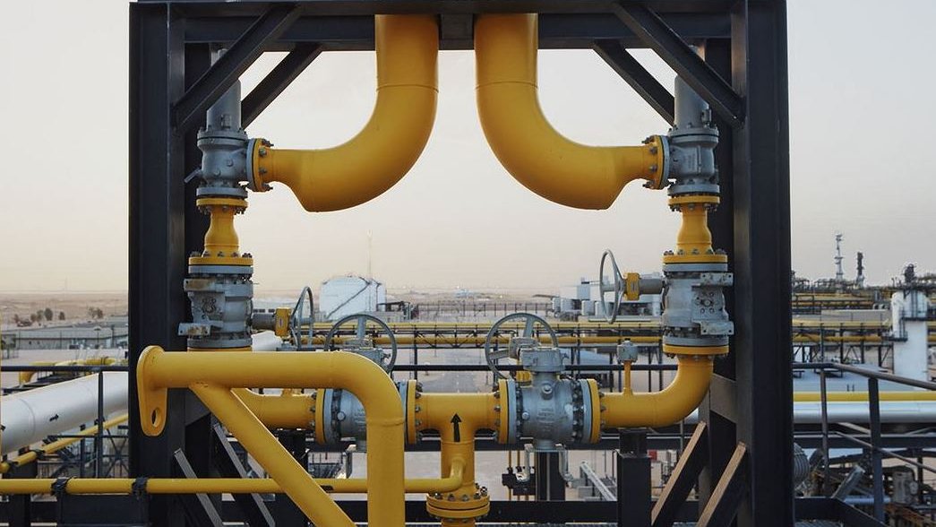 La imagen muestra una estructura industrial, probablemente de una planta de procesamiento de petróleo o gas. Hay varias tuberías de color amarillo que se conectan entre sí, junto con válvulas y otros elementos metálicos. El fondo sugiere un paisaje industrial con tanques y equipos, lo que indica un entorno de trabajo relacionado con la energía o la petroquímica. La escena parece ser al aire libre, bajo un cielo claro.
