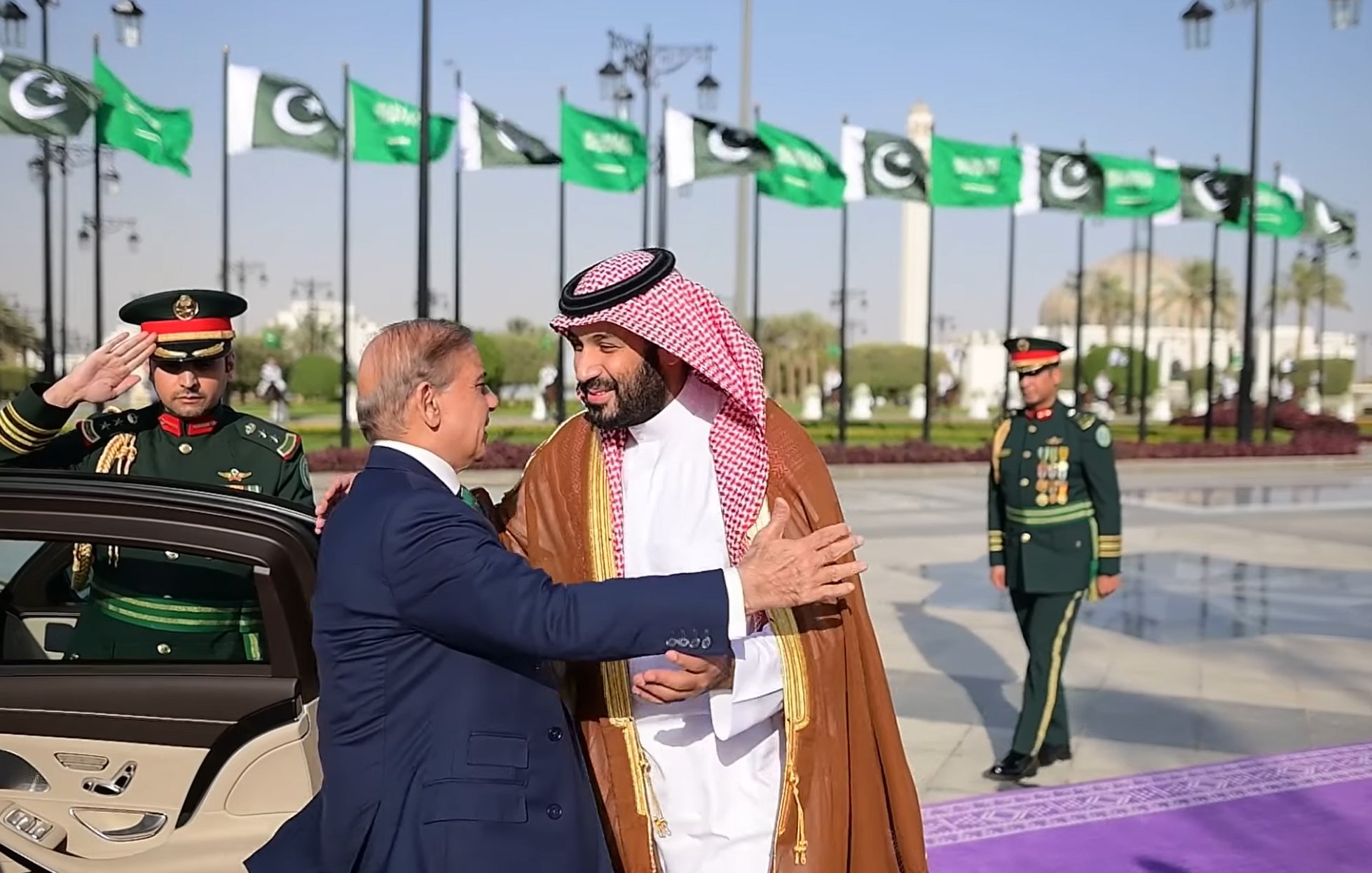 A formal greeting between two leaders, with guards and flags in the background.