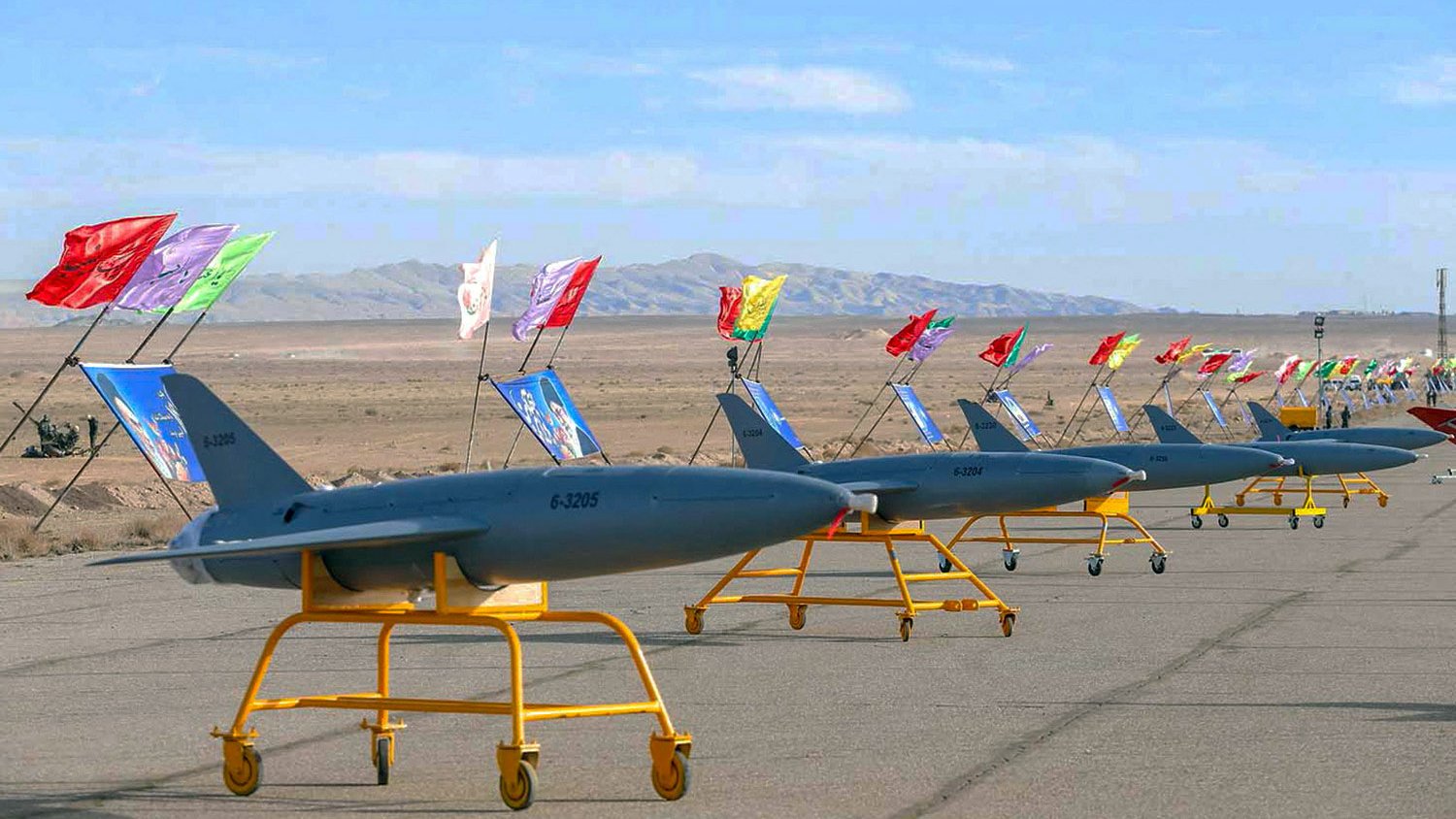 La imagen muestra una fila de drones en una pista desértica. Estos drones están montados sobre soportes amarillos y llevan banderas de diferentes colores. Al fondo, se puede ver un paisaje montañoso y un cielo despejado. La escena parece estar relacionada con una exhibición o un evento militar.