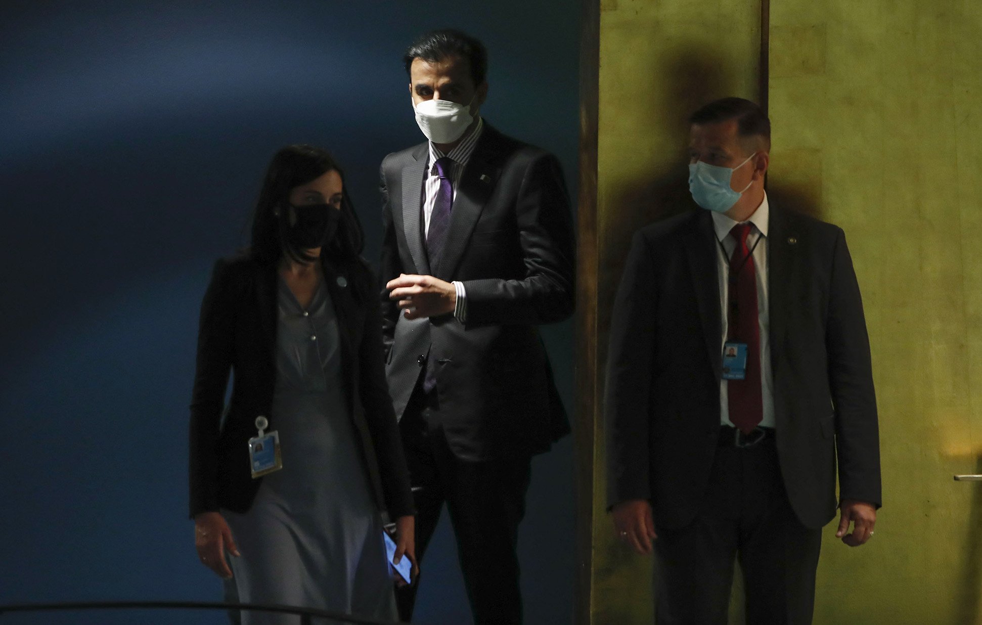 La imagen muestra a tres personas en un entorno que parece ser un edificio oficial, probablemente en una reunión o evento. Dos de ellos llevan mascarillas y lucen formales, acomodándose a un ambiente institucional. Una mujer está a la izquierda, con un vestido gris y una identificación, mientras que un hombre en el centro, vestido de traje, parece estar conversando o aguardando. A la derecha, hay otro hombre también en traje, observando. El fondo tiene colores oscuros y brillantes, sugiriendo una atmósfera seria y profesional.