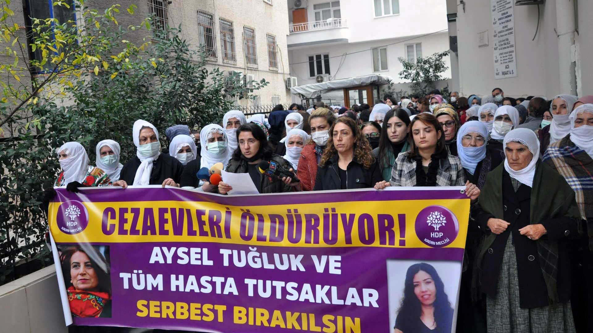 En la imagen, un grupo de personas se encuentra reunido en una manifestación. Hay mujeres con pañuelos en la cabeza, y algunas de ellas sostienen pancartas que expresan mensajes en apoyo a Aysel Tuğluk y a otros prisioneros enfermos. El ambiente parece ser de protesta, con un enfoque en la liberación de detenidos. Se observa una edificación al fondo, que sugiere que la manifestación se lleva a cabo frente a un edificio público o institucional.