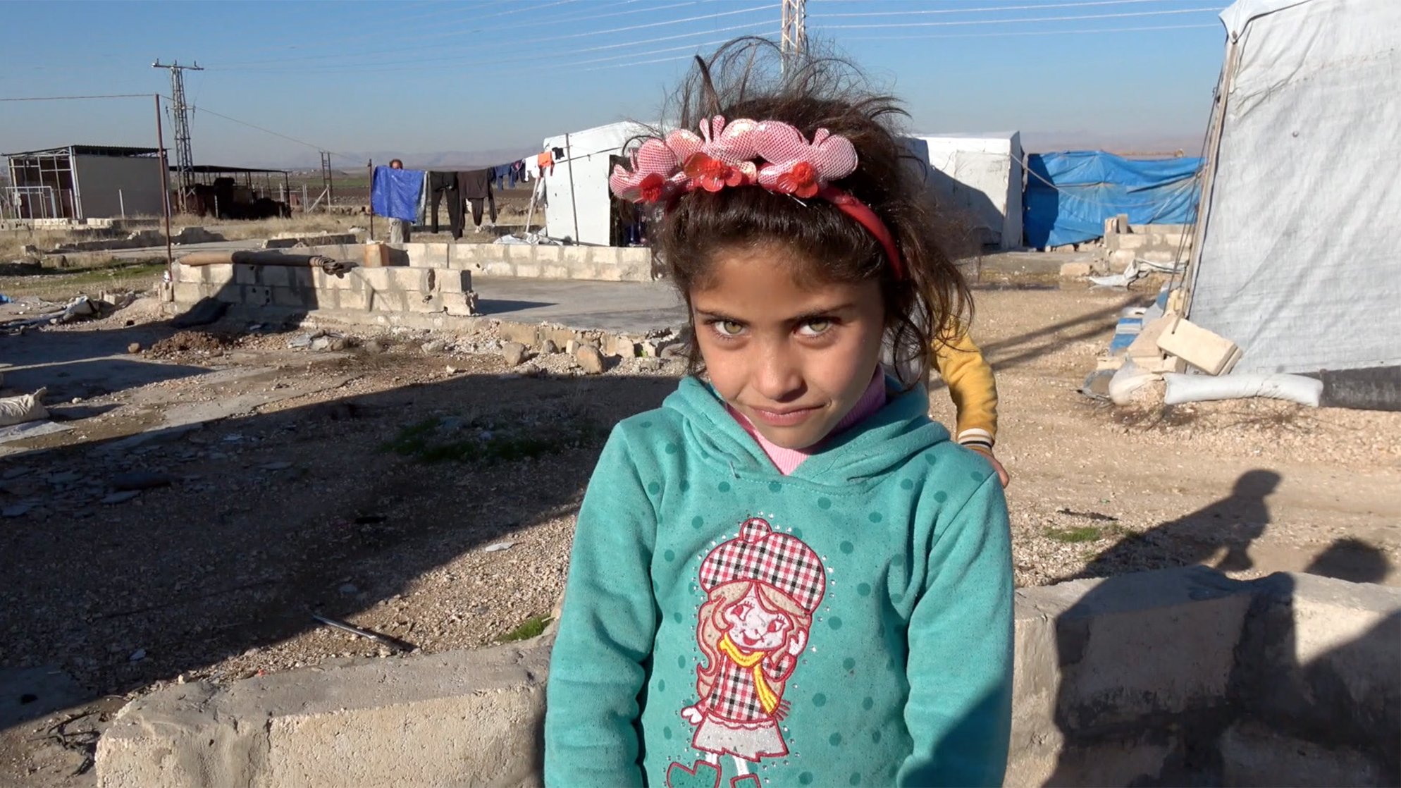 La imagen muestra a una niña pequeña en un campo de refugiados. Ella tiene largas pestañas y ojos claros, y lleva una sudadera de color azul con un diseño en la parte delantera. En su cabeza, tiene una diadema decorada con flores. El entorno es árido, con estructuras de refugio improvisadas y un cielo despejado. La expresión de la niña es curiosa y ligeramente sonriente.