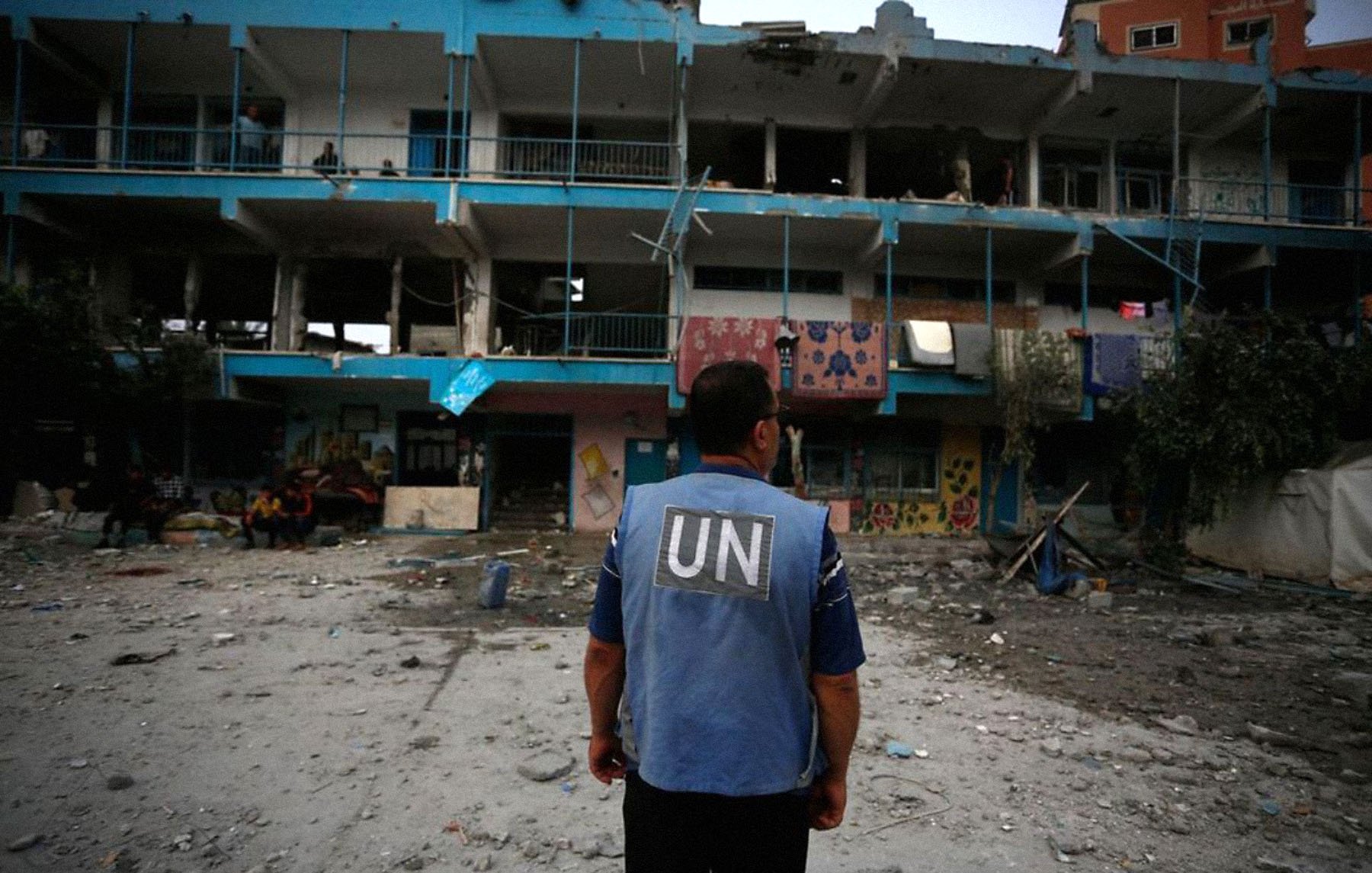 The image depicts a person wearing a blue vest labeled "UN," standing in front of a damaged building. The structure shows signs of destruction, with broken windows and debris scattered around the ground. There are individuals visible in the background, and the overall scene conveys a somber atmosphere, likely in a conflict or disaster-stricken area.