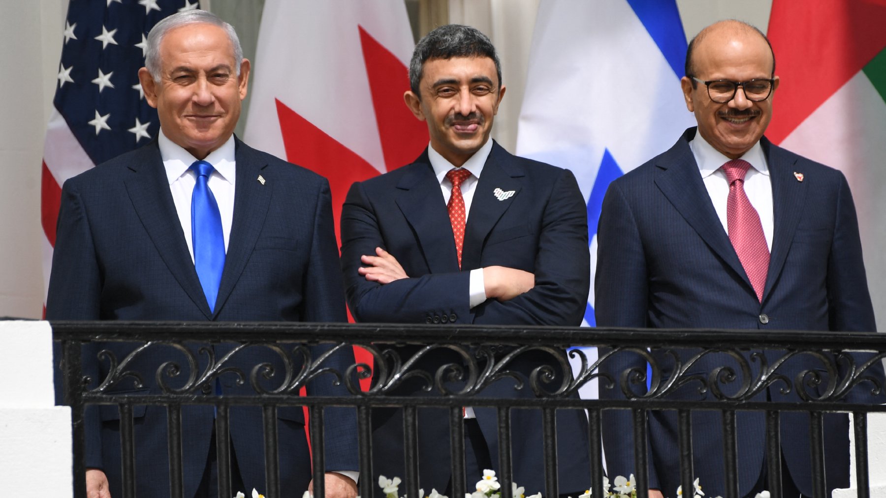 En la imagen se pueden ver a tres hombres de pie en una terraza, sonriendo. Al fondo hay banderas de Estados Unidos, Canadá, Israel y otros países. Los hombres están vestidos con trajes formales: el de la izquierda lleva una corbata azul, el del centro tiene una corbata roja y el de la derecha lleva una corbata a rayas rojas y azules. En la barandilla hay flores blancas. La atmósfera parece ser positiva y amistosa.