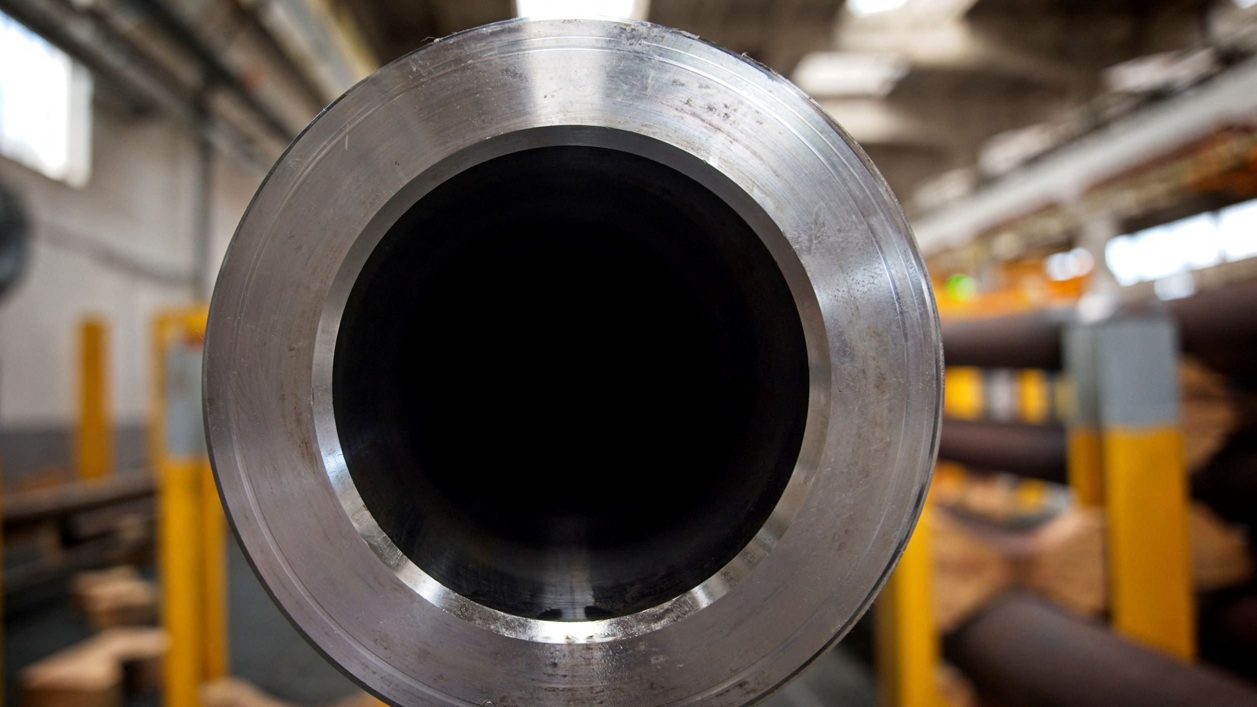 La imagen muestra el extremo de un tubo de metal, donde se puede ver su interior oscuro y liso. El tubo tiene un acabado brillante en su parte exterior, y el entorno sugiere un ambiente industrial, con otros tubos y estructuras a lo lejos, pero enfocados en primero plano. La iluminación es suave y resalta los detalles del material.