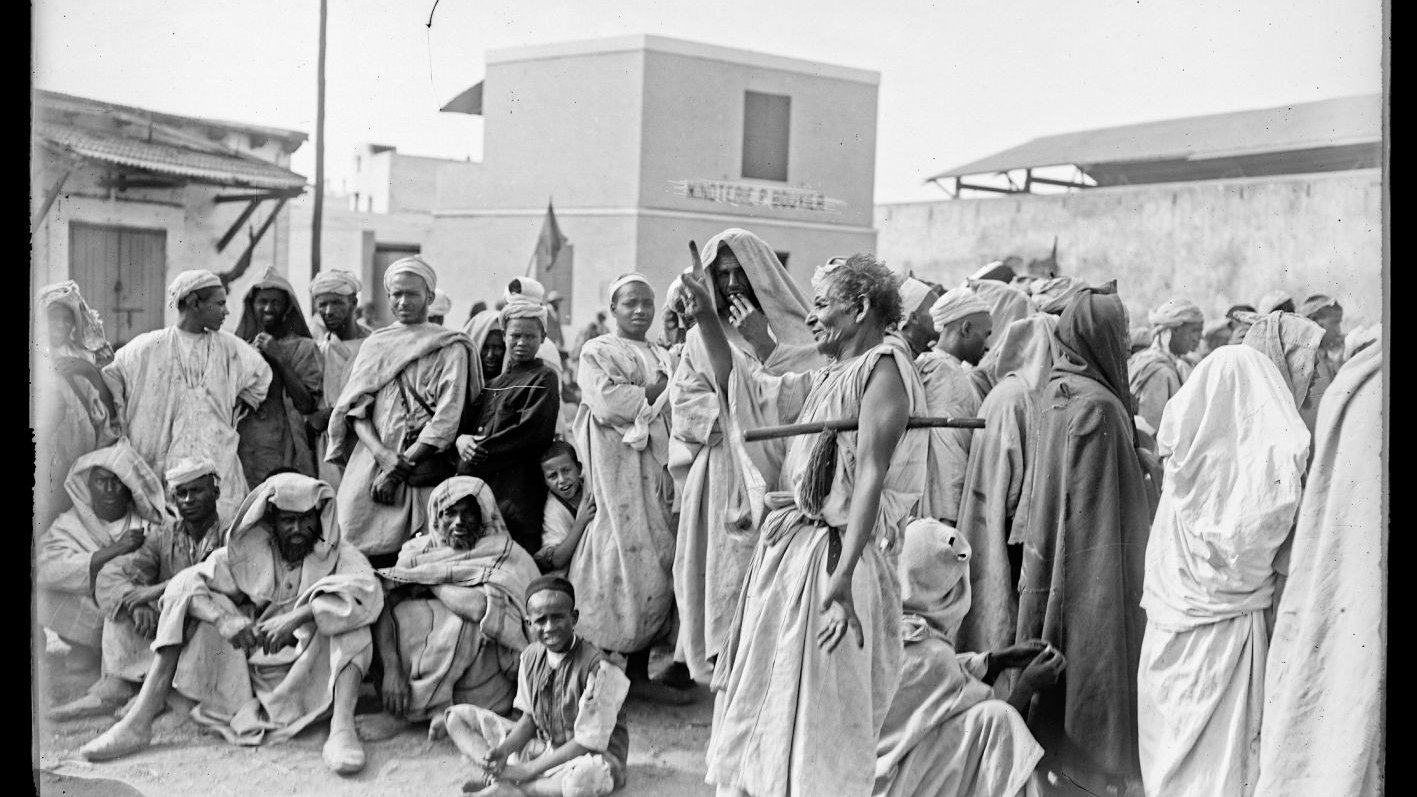 La imagen muestra a un grupo de personas en un entorno que parece ser un mercado o una reunión al aire libre. La mayoría de las personas están vestidas con ropas tradicionales, muchas de las cuales son de colores claros y están cubiertas con prendas que podrían indicar el clima cálido de la región. Hay un enfoque particular en una figura central que parece estar gesticulando o hablando, rodeada de un público atento. El ambiente es de interacción social y hay una variedad de expresiones en los rostros de las personas, que van desde la curiosidad hasta la concentración. El fondo muestra edificaciones simples, lo que sugiere un contexto urbano o rural.