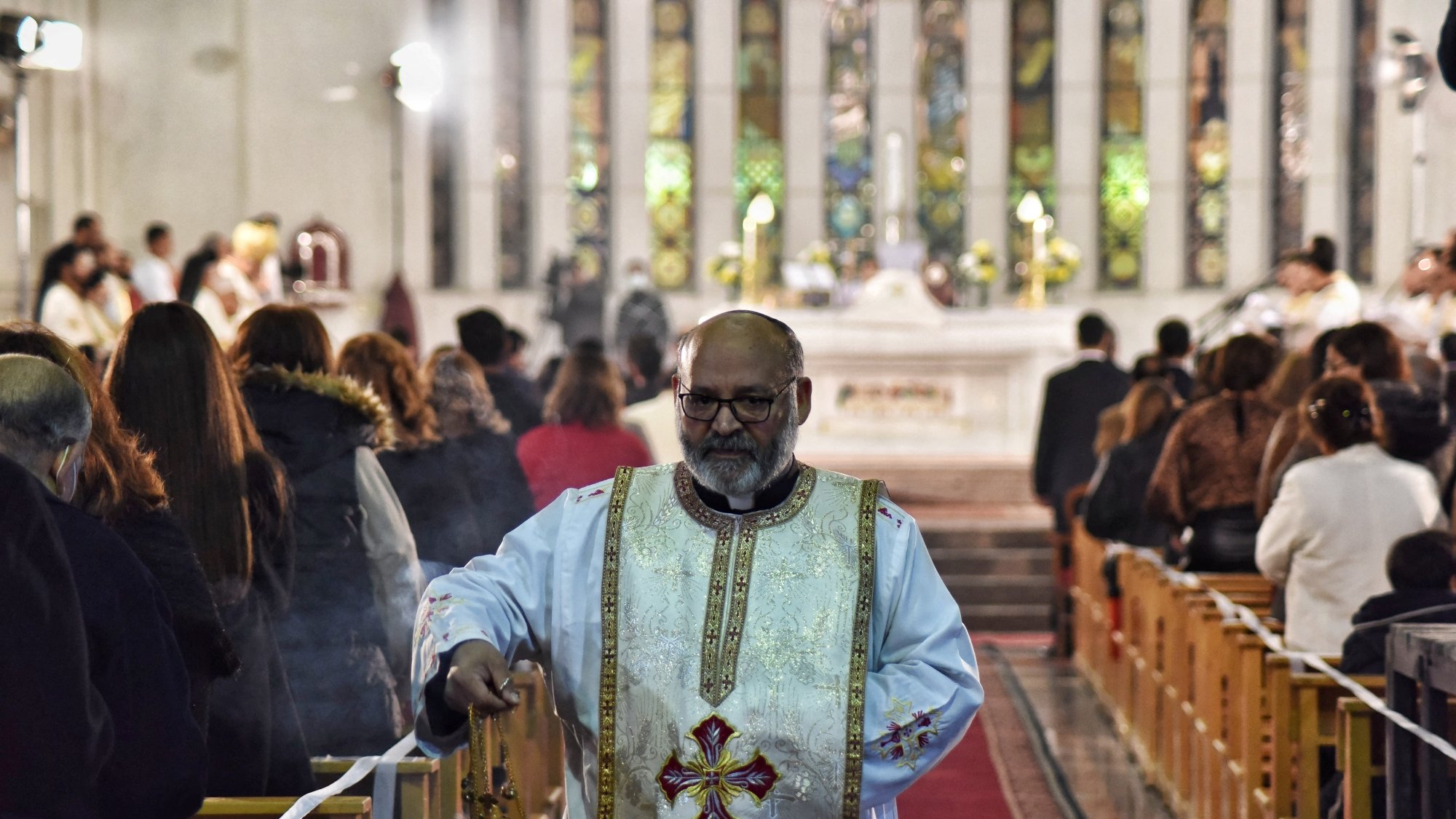 A priest in ornate vestments walks down an aisle in a church, surrounded by worshippers.