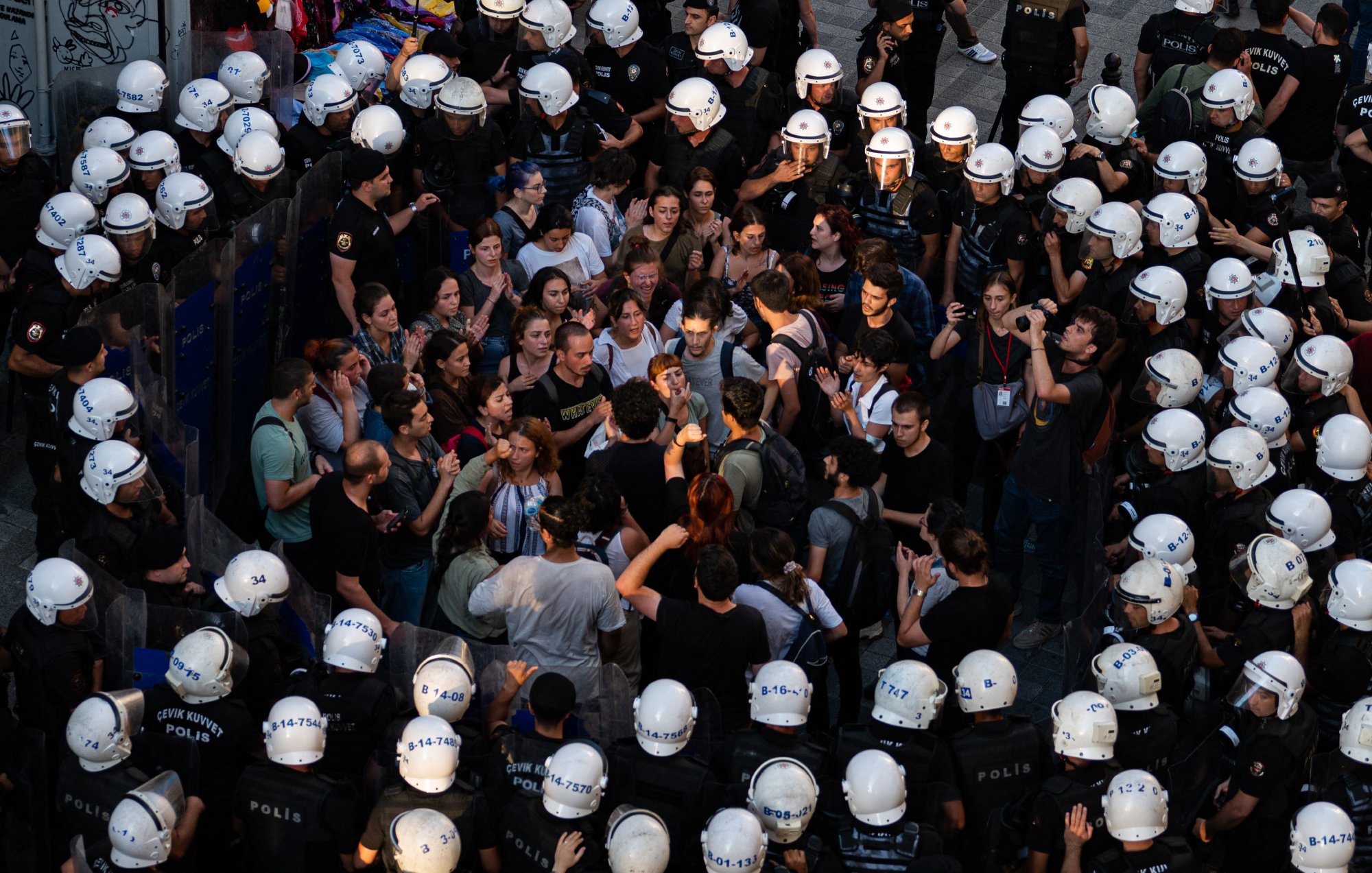 La imagen muestra una multitud de personas rodeadas por un grupo de policías. Los manifestantes parecen estar en una discusión o protesta, expresando sus opiniones entre ellos. Los policías, con cascos y uniformes, forman un cordón alrededor de la multitud, lo que sugiere una situación tensa. La escena parece tener lugar en un ambiente urbano, posiblemente durante una manifestación o conflicto social. La iluminación sugiere que puede ser al atardecer o al final del día.