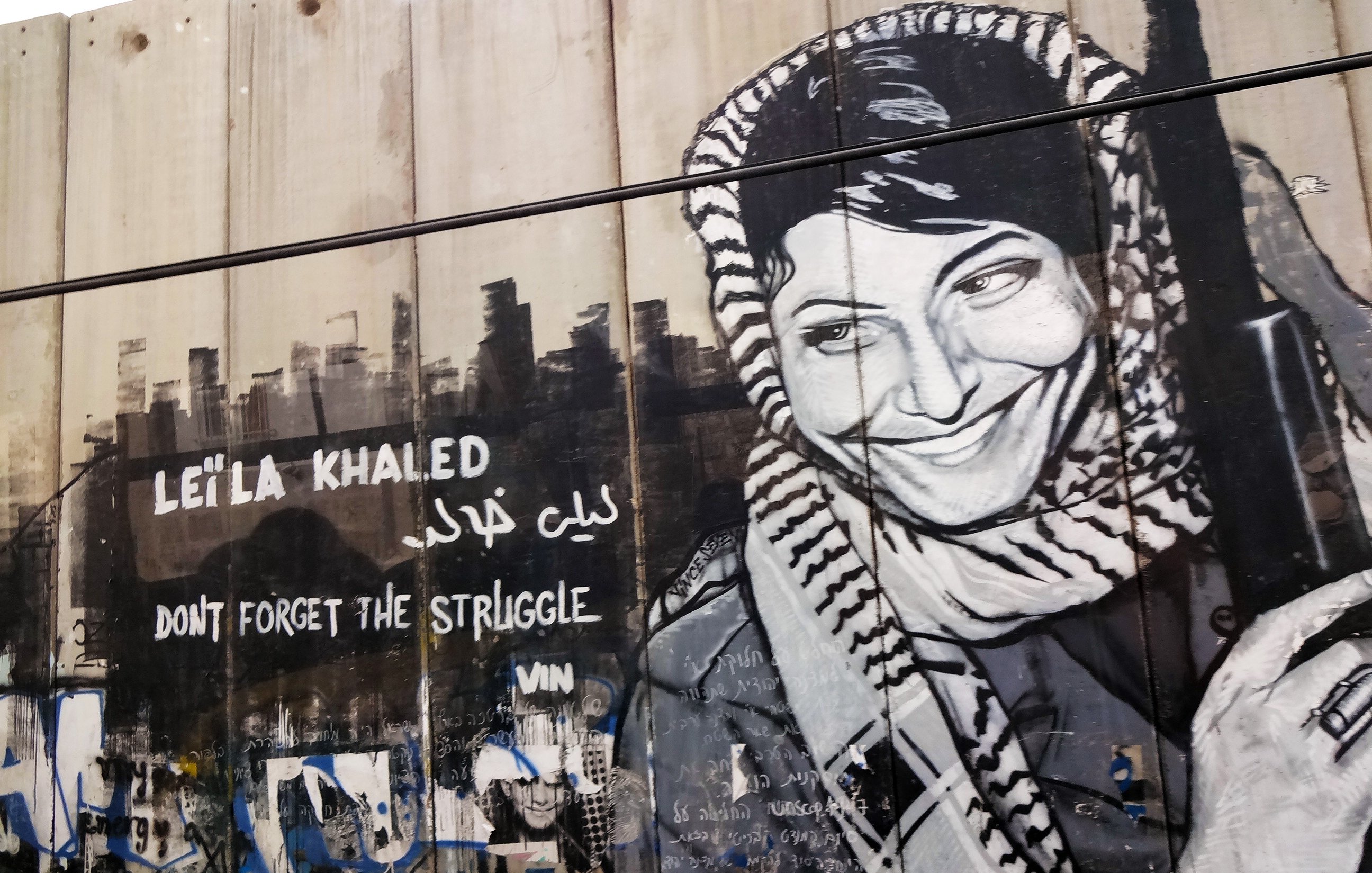 La imagen muestra un mural pintado en una pared, con un retrato en blanco y negro de una mujer que sonríe, vistiendo un keffiyeh. Junto a su imagen, hay texto en inglés que dice "DON'T FORGET THE STRUGGLE" y en árabe el nombre "ليلى خالد" (Leila Khaled). Este mural parece rendir homenaje a la figura de Leila Khaled, conocida por su activismo político y su papel en la lucha palestina. El fondo de la pared tiene grafitis y otros símbolos, lo que añade contexto urbano y de resistencia.