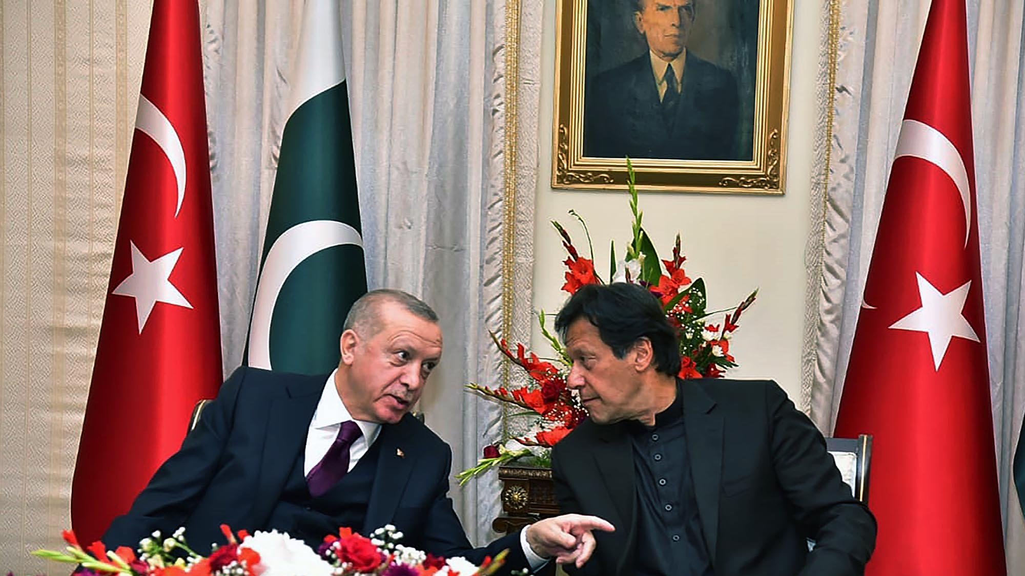 En la imagen se pueden ver a dos hombres conversando en un ambiente formal, rodeados de banderas de Turquía y Pakistán. Hay un arreglo floral colorido en la mesa, y detrás de ellos se encuentra un retrato en la pared. La escena transmite un ambiente de diplomacia y colaboración entre los dos países.
