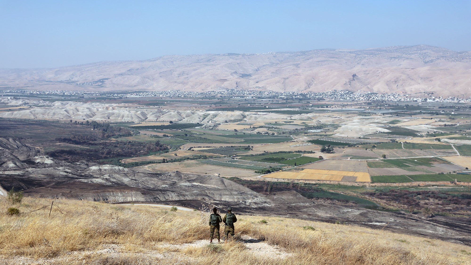 La imagen muestra un paisaje vasto y montañoso, con colinas y valles amplios. En primer plano, se pueden ver dos personas de pie, posiblemente soldados, observando el paisaje desde una cima. El área está cubierta de hierba seca y destaca un amplio valle con campos de cultivo en diferentes tonos de verde y amarillo. En el fondo, se aprecian montañas que se extienden hasta el horizonte bajo un cielo despejado.