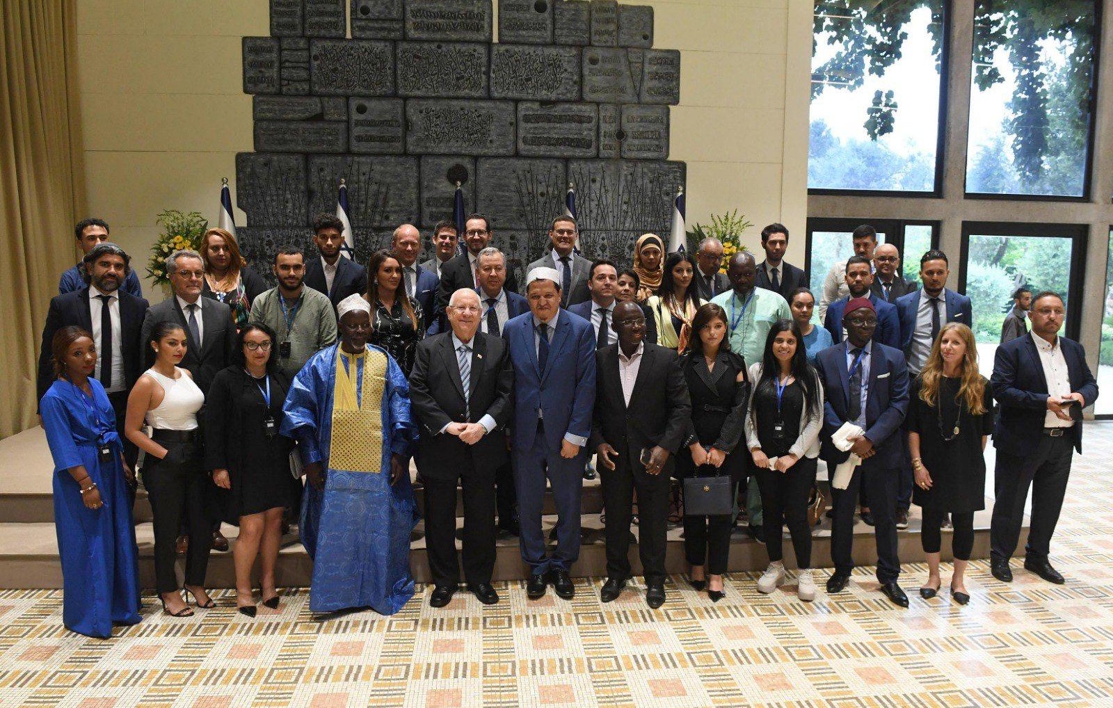 La imagen muestra a un grupo de personas posando juntas en un espacio interior bien iluminado. Al fondo, se pueden ver banderas y una pared decorativa. El grupo incluye hombres y mujeres de diversas apariencias, algunos con trajes formales y otros con atuendos tradicionales. Todos están sonriendo y parecen reunidos para una ocasión especial, posiblemente un evento diplomático o cultural. La disposición del grupo sugiere una atmósfera de cooperación y celebración.
