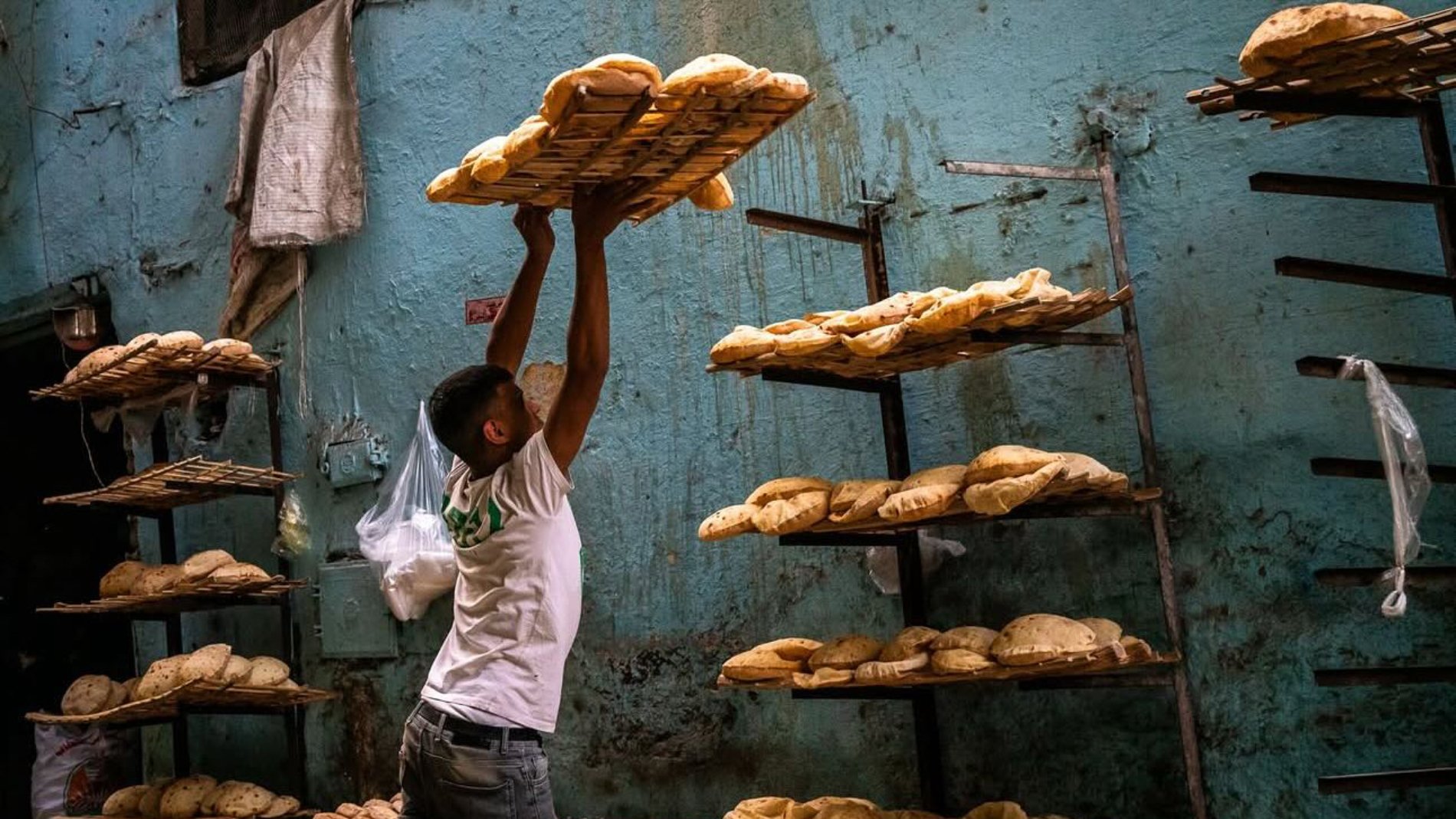 Un hombre levanta canastas con pan en un ambiente de panadería. Las paredes son azules y desgastadas.