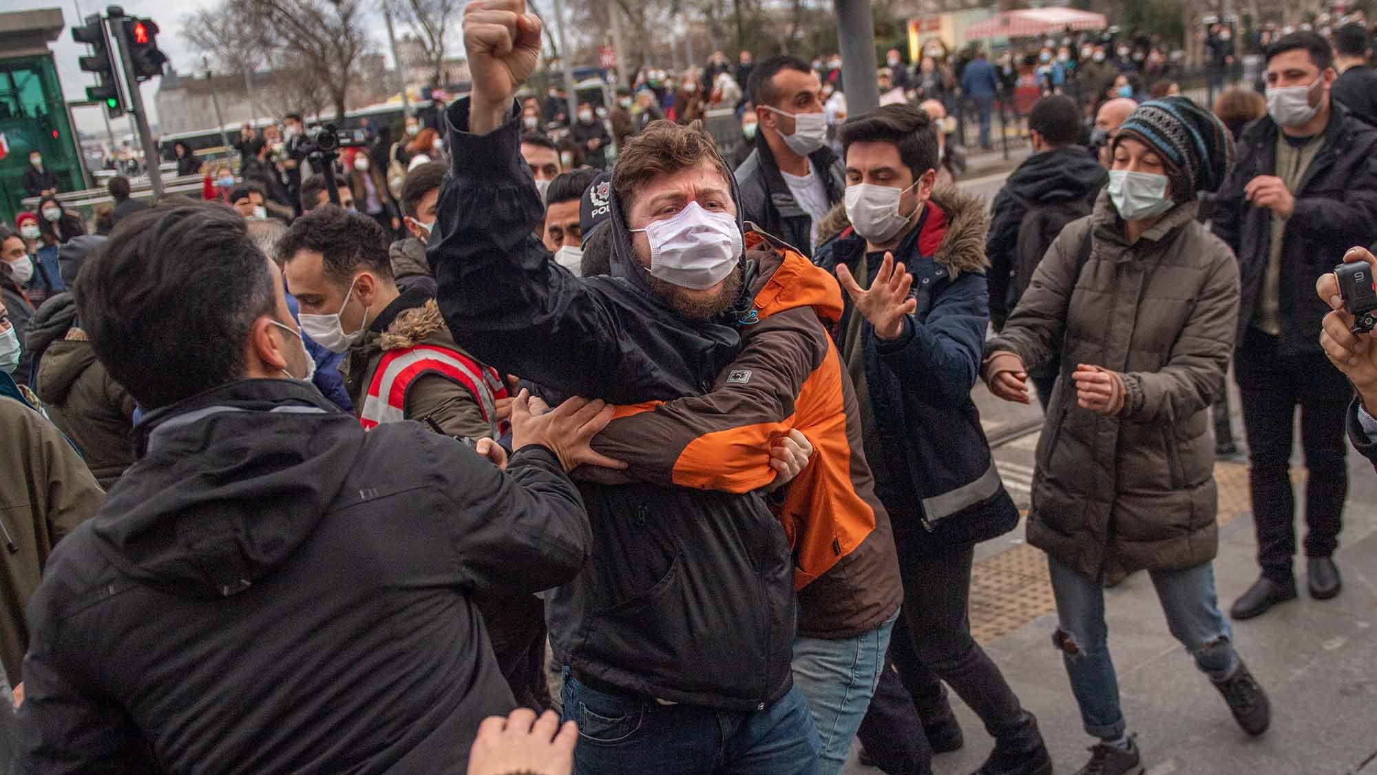 La imagen muestra a un grupo de personas en medio de una situación tensa. Se puede ver a un individuo en el centro, levantando el puño mientras es sujetado por otro, aparentemente en una escena de protesta o manifestación. La mayoría de las personas en la imagen llevan mascarillas. El ambiente parece ser urbano, con un semáforo y algunos edificios de fondo. La expresión de los individuos sugiere emociones intensas, probablemente relacionadas con el conflicto o la agitación social.
