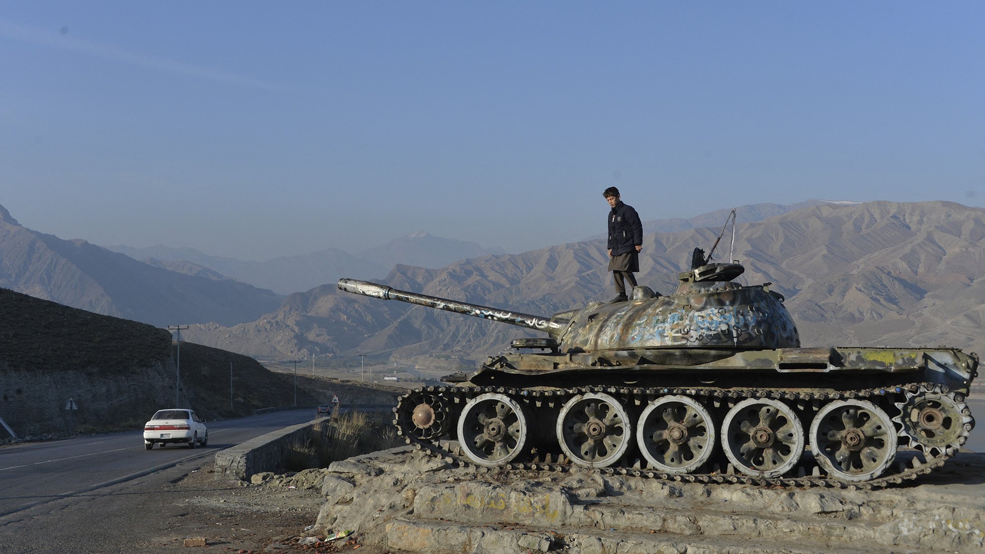 La imagen muestra a un joven de pie sobre un tanque de guerra, que parece estar fuera de uso. El tanque tiene un acabado desgastado y está cubierto de óxido y pintura descascarada. Al fondo, se pueden ver montañas cubiertas de vegetación y un cielo despejado. A un lado, hay una carretera donde se observa un automóvil blanco en movimiento. La escena transmite una sensación de desolación y el contraste entre la guerra y la paz.