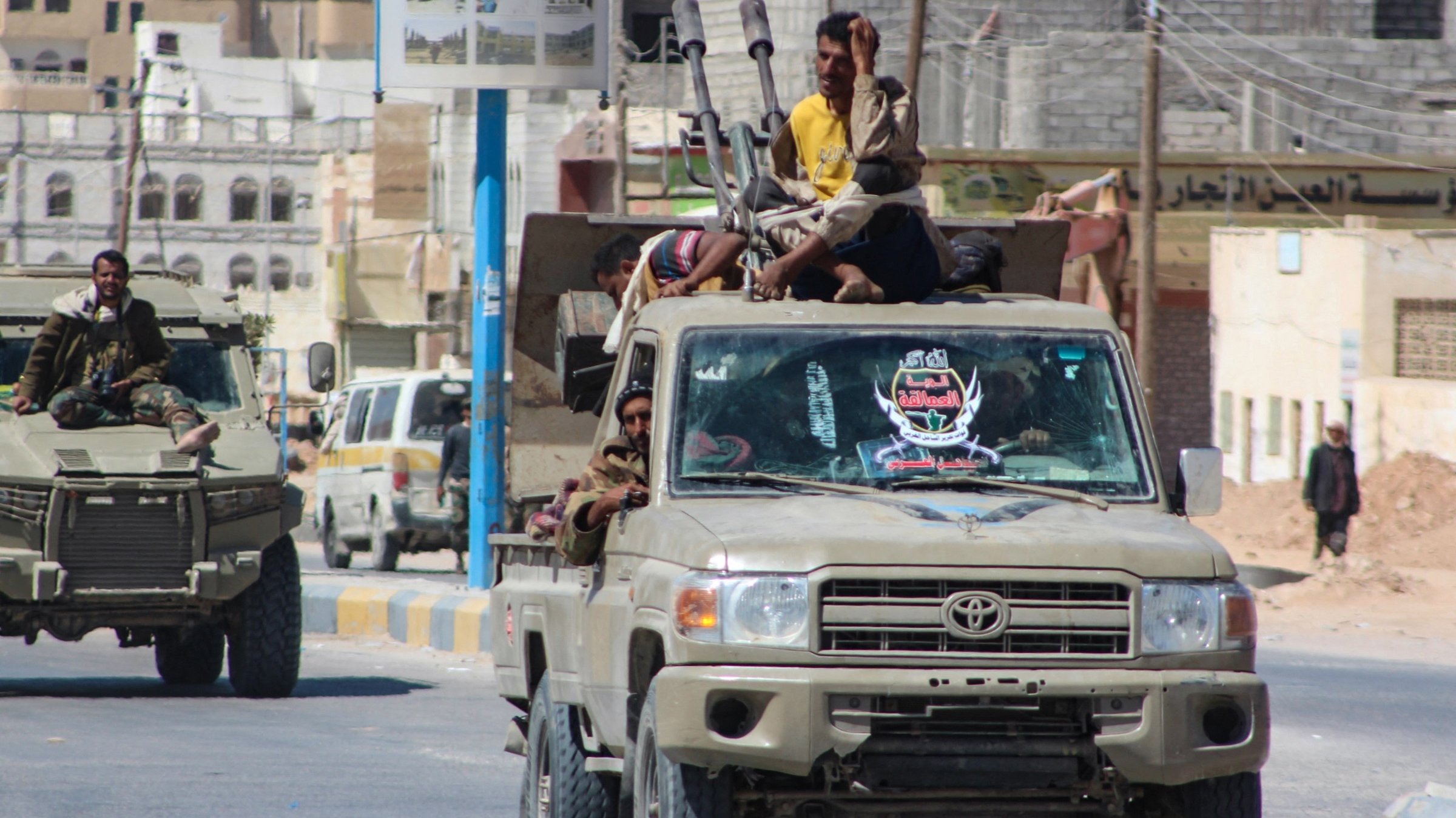 La imagen muestra un convoy de vehículos militares, principalmente camiones, en un ambiente urbano. En uno de los camiones, varias personas están sentadas y de pie en la parte trasera. Algunos están armados y parecen estar en posición de vigilancia. Los vehículos tienen un color beige o khaki, típico de vehículos militares. En el fondo, se pueden ver edificios y una carretera, lo que indica que están en una zona urbana. La escena transmite una sensación de actividad militar en la calle.