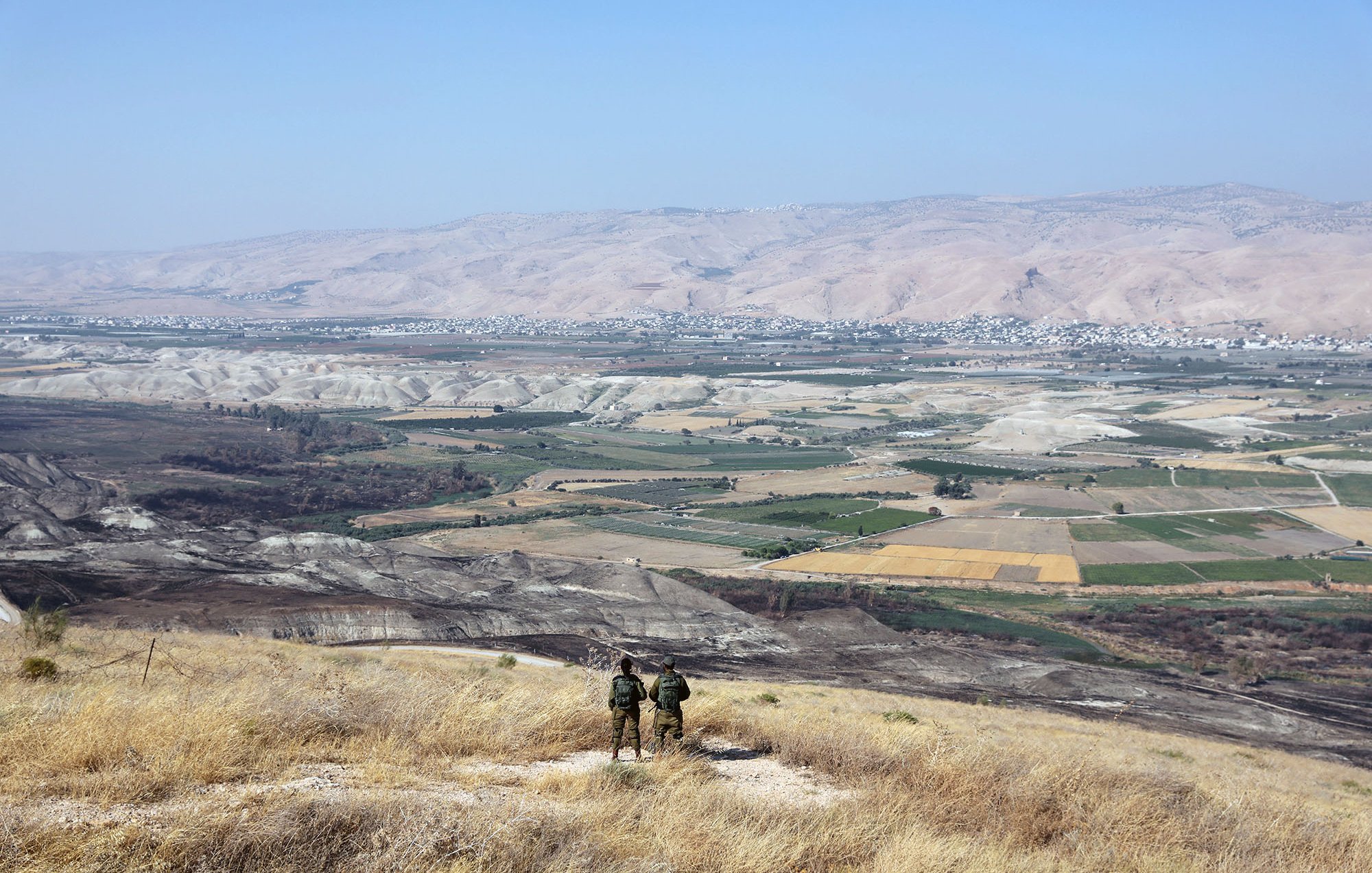 La imagen muestra un paisaje vasto y montañoso, con colinas y valles amplios. En primer plano, se pueden ver dos personas de pie, posiblemente soldados, observando el paisaje desde una cima. El área está cubierta de hierba seca y destaca un amplio valle con campos de cultivo en diferentes tonos de verde y amarillo. En el fondo, se aprecian montañas que se extienden hasta el horizonte bajo un cielo despejado.