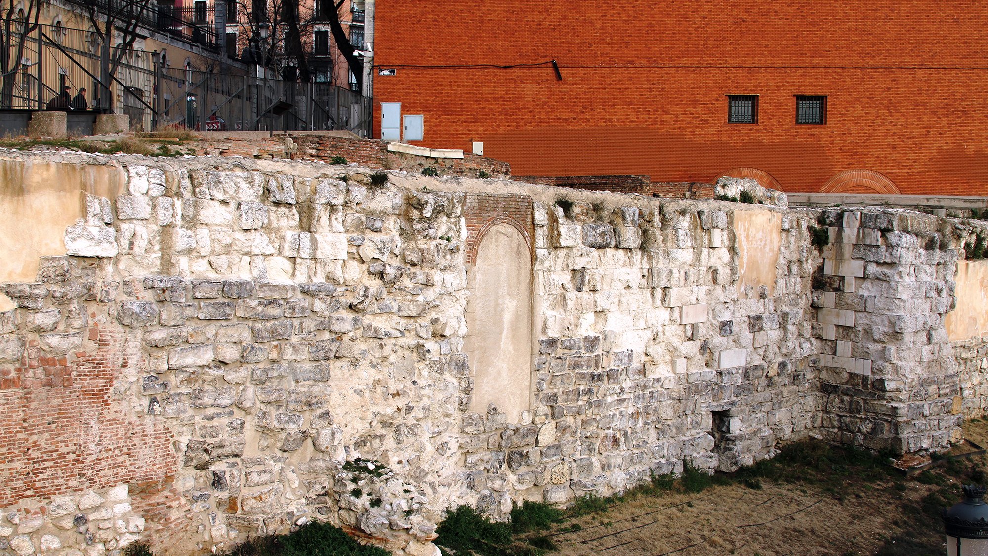 La imagen muestra una pared de piedra antigua, probablemente parte de una estructura histórica. La pared está compuesta de grandes bloques de piedra, algunos de los cuales están desgastados o dañados. En el centro, hay un espacio vacío en la pared que sugiere la existencia previa de una puerta o ventana. Al fondo, se puede ver un edificio de ladrillo rojo y un área de césped seco. Hay algunos restos de vegetación en la base de la pared, lo que añade un toque de naturaleza al entorno urbano.