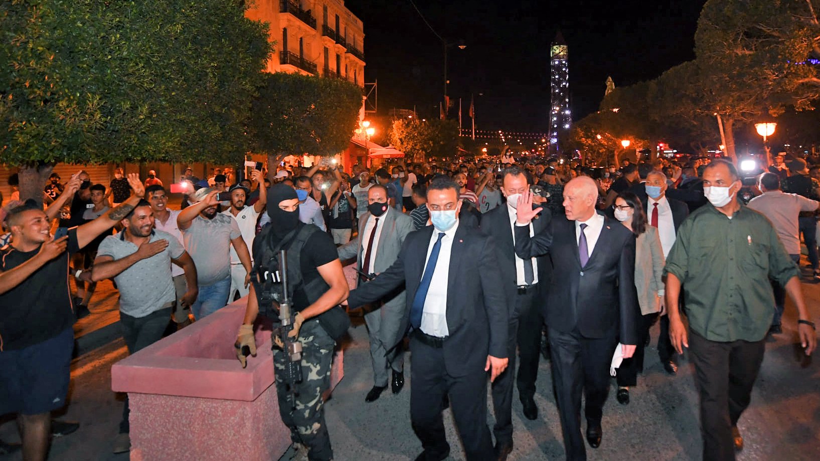 La imagen muestra a un grupo de personas en una calle durante la noche. En el centro, hay un hombre vestido con traje y corbata, escoltado por un guardia de seguridad con una máscara y uniforme. Alrededor, se ven varios manifestantes levantando las manos, algunos parecen estar enérgicos o en protesta. La iluminación de la escena proviene de faroles y luces en el entorno, creando un ambiente animado y posiblemente tenso. Los árboles y edificios en el fondo sugieren que se trata de una zona urbana.