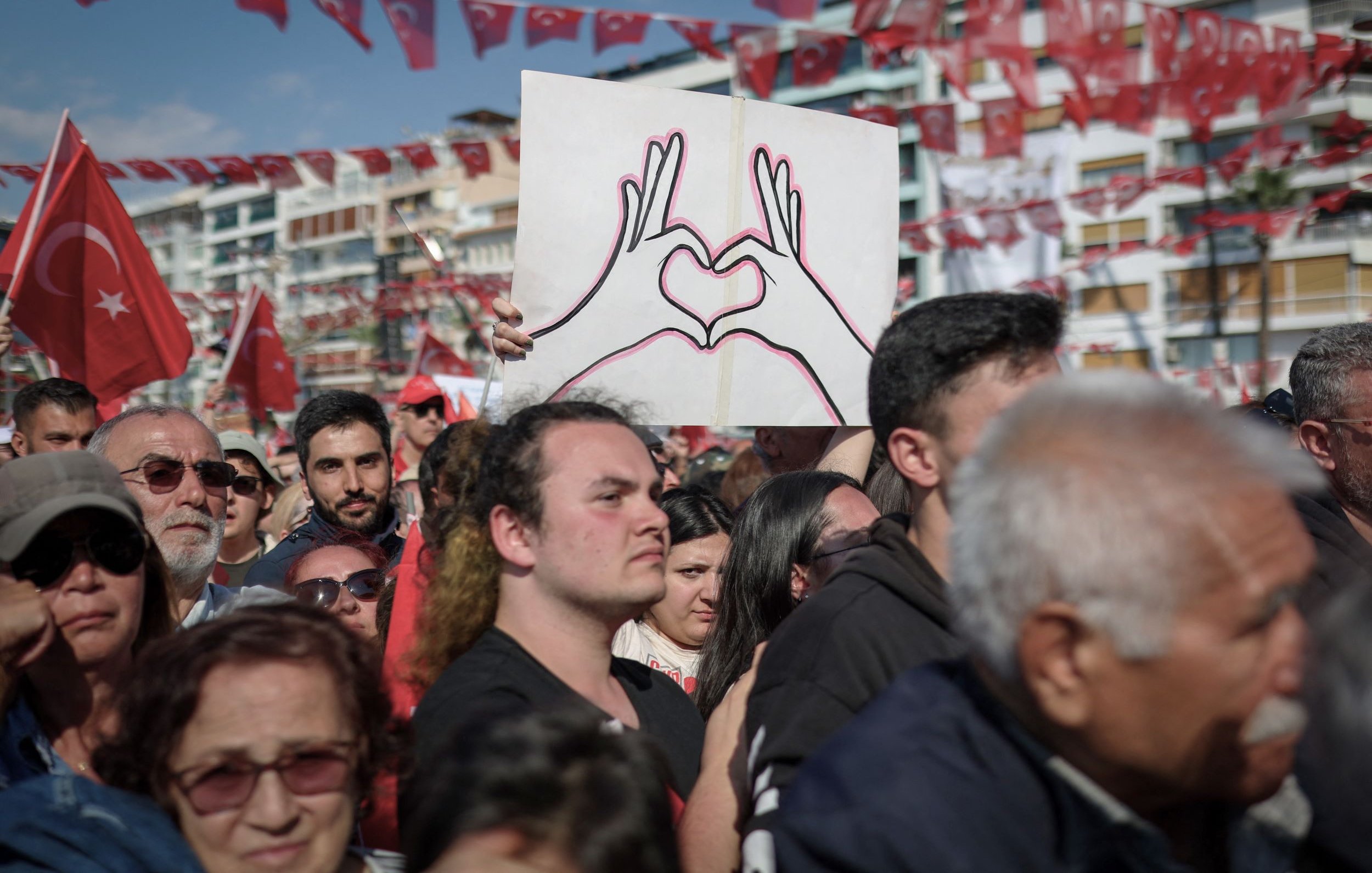 La imagen muestra una multitud reunida en un evento, posiblemente una manifestación o celebración. En primer plano, hay un hombre que sostiene un cartel en forma de corazón, dibujado con las manos. La multitud parece diversa y está vestida con ropa casual. Al fondo, hay banderas rojas con la insignia de Turquía, lo que sugiere un contexto nacional. La atmósfera parece ser de unidad y orgullo colectivo.