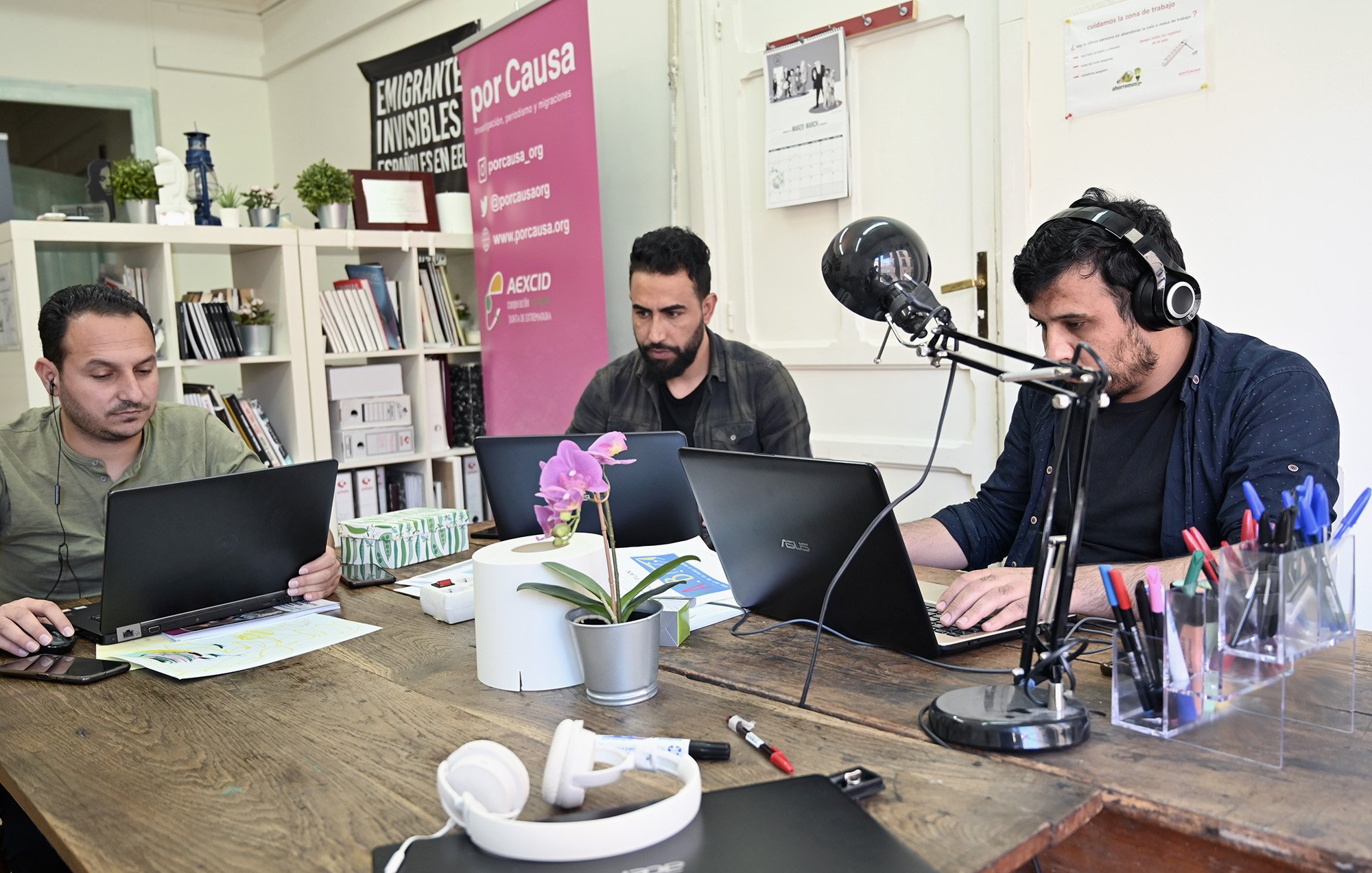 En la imagen se pueden ver a tres hombres sentados en una mesa de trabajo. Cada uno tiene un portátil frente a él y parece que están concentrados en sus tareas. La mesa está decorada con una planta en una maceta y hay auriculares sobre la superficie. En el fondo, hay una pared con algunos carteles, incluyendo uno que dice "Emigrantes Invisibles". También hay estantes con libros y otros materiales. La atmósfera sugiere un ambiente de trabajo colaborativo y dinámico.