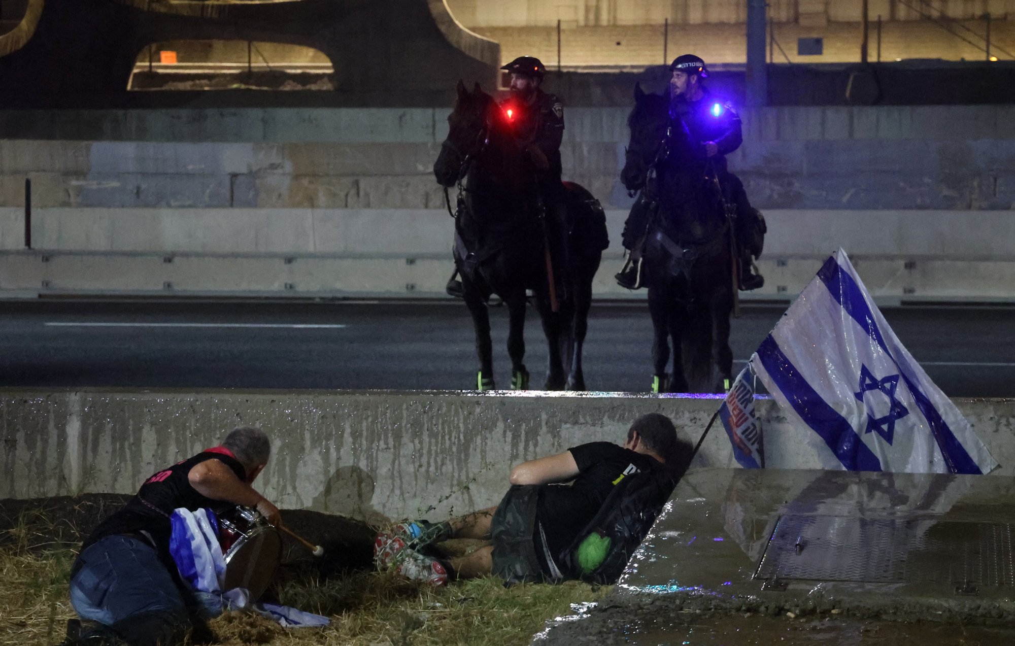 La imagen muestra a un par de oficiales montados sobre caballos, con luces rojas y azules, vigilando una situación que parece estar relacionada con una protesta. En el primer plano, hay dos personas en el suelo, una de las cuales sostiene una bandera de Israel. La escena parece indicar una tensión o un conflicto en el contexto de la protesta, con un ambiente urbano de fondo, probablemente en una carretera. La iluminación y la posición de los agentes reflejan la seriedad de la situación.