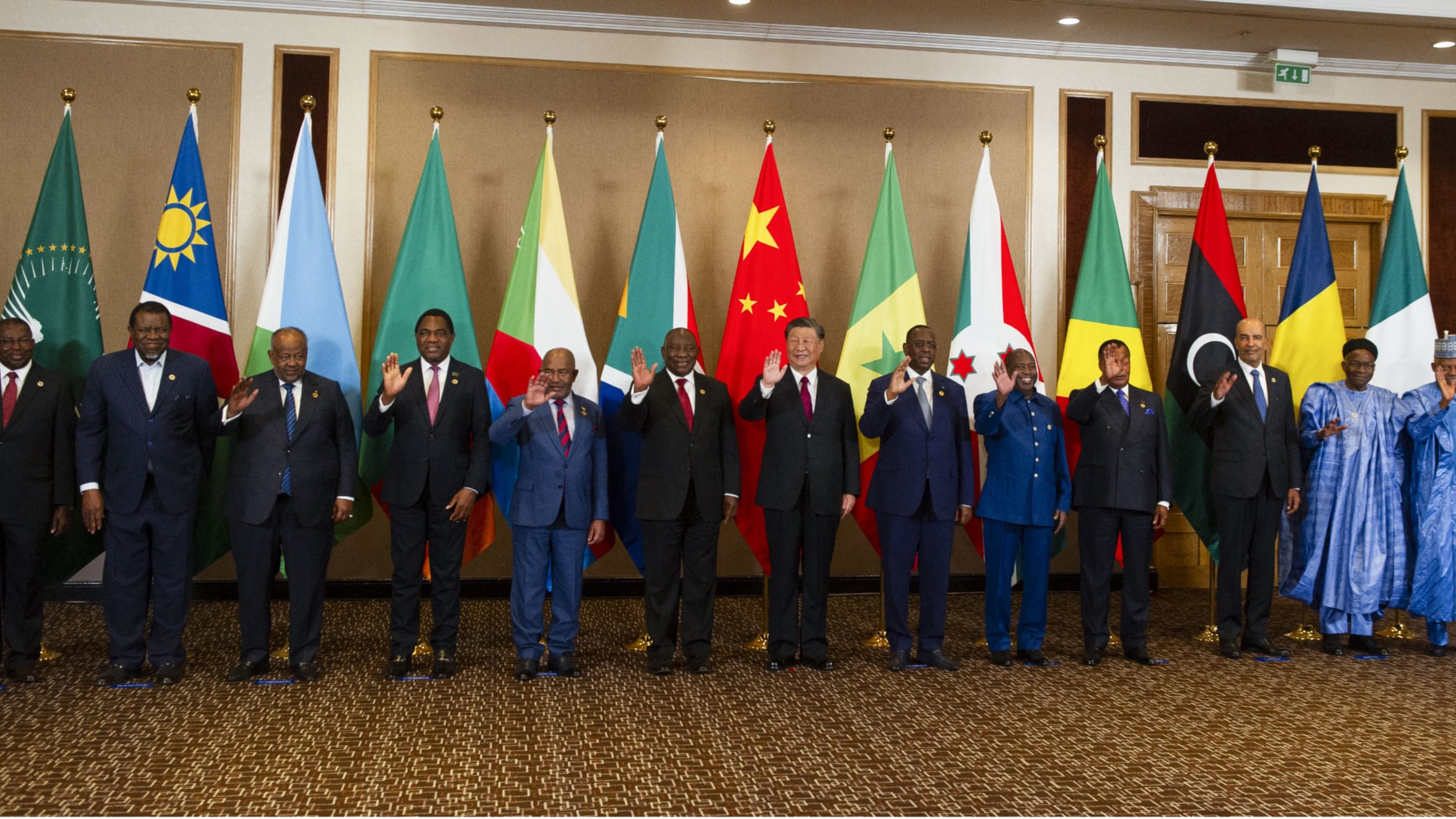 La imagen muestra a un grupo de líderes y representantes de diferentes países alineados en una sala. Detrás de ellos hay banderas de varias naciones, incluidos países africanos y de otras partes del mundo. Todos los individuos parecen estar posando para una fotografía, algunos levantando la mano en señal de saludo. La decoración del lugar es elegante, con un ambiente formal que refleja la importancia del evento.