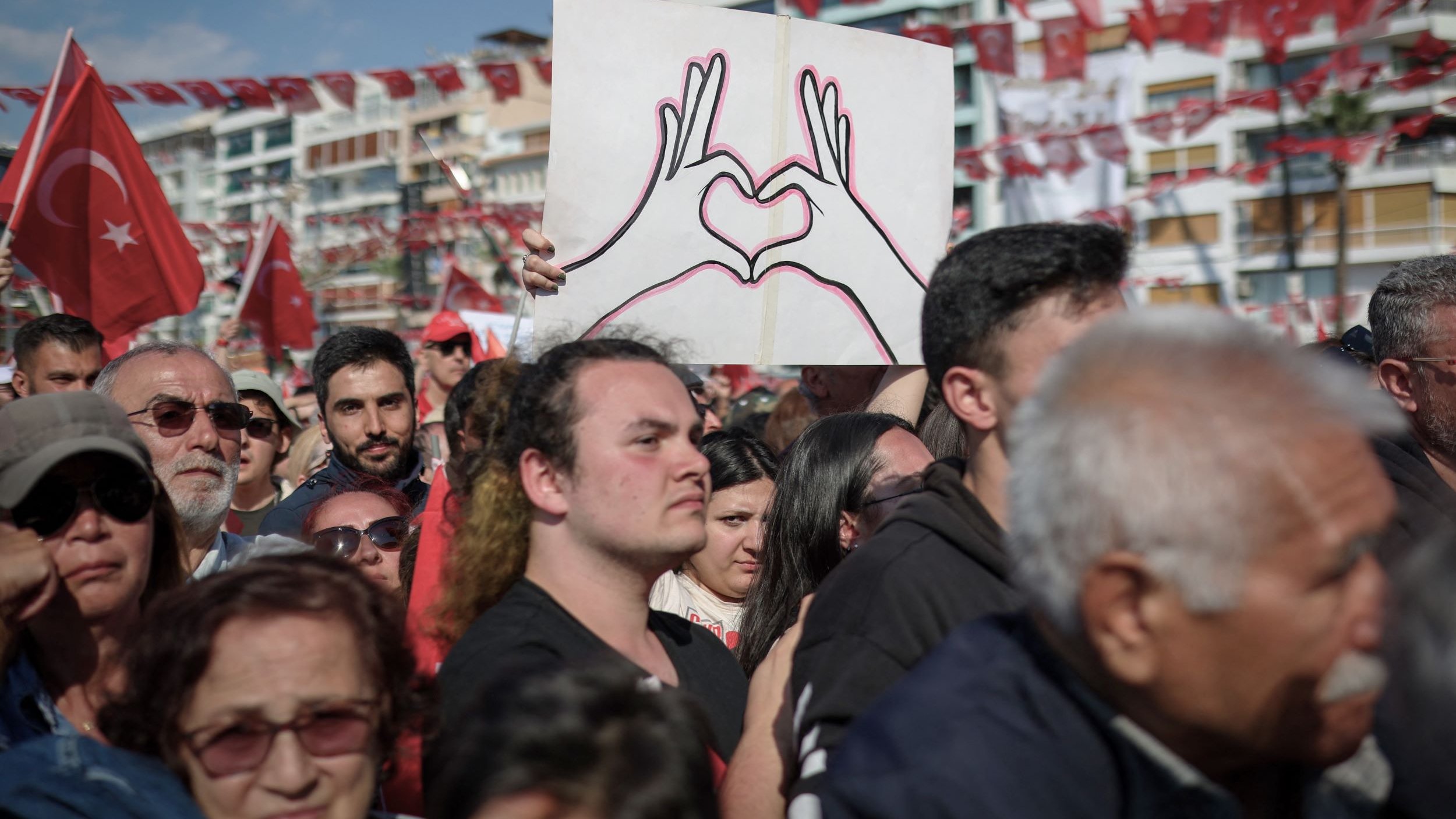 La imagen muestra una multitud reunida en un evento, posiblemente una manifestación o celebración. En primer plano, hay un hombre que sostiene un cartel en forma de corazón, dibujado con las manos. La multitud parece diversa y está vestida con ropa casual. Al fondo, hay banderas rojas con la insignia de Turquía, lo que sugiere un contexto nacional. La atmósfera parece ser de unidad y orgullo colectivo.