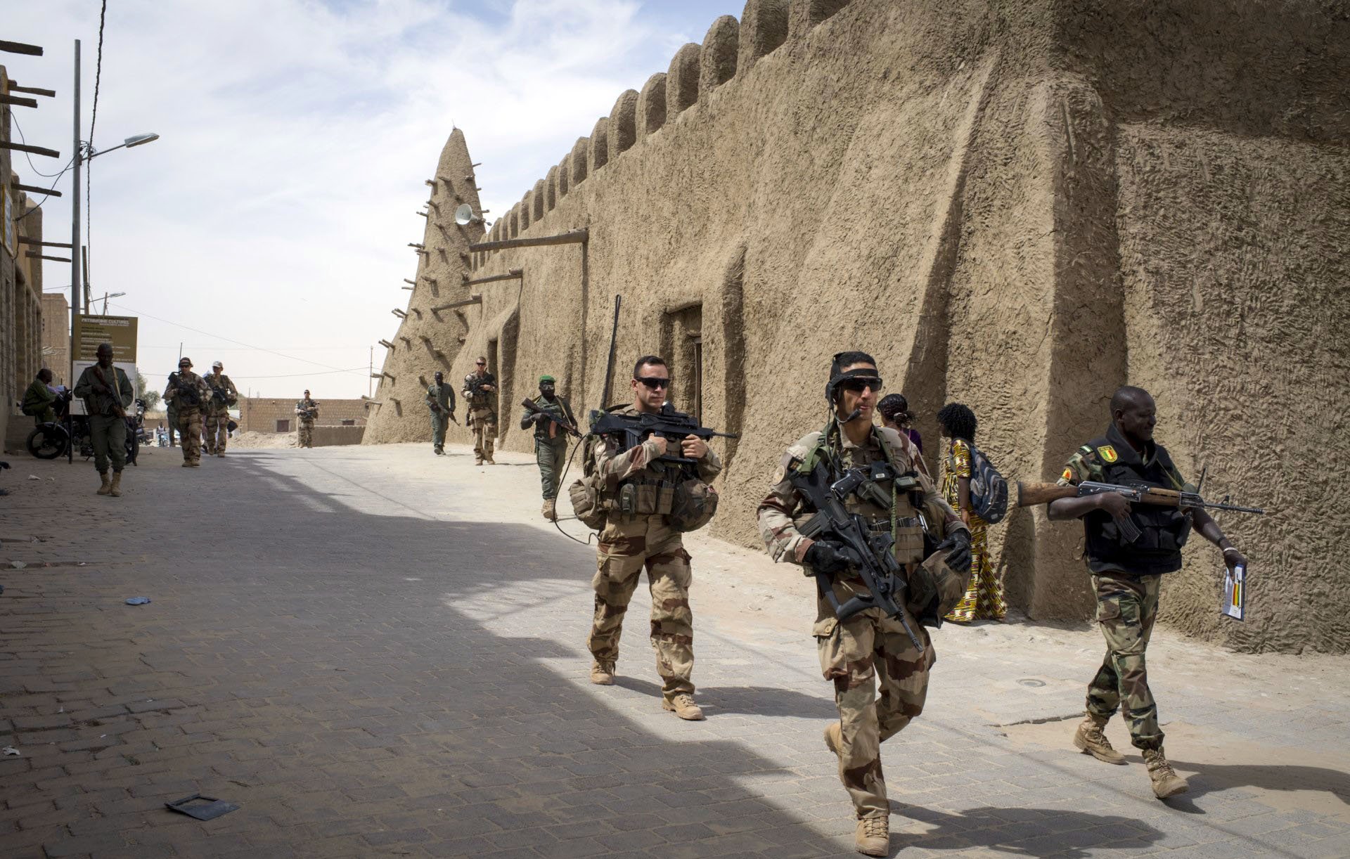 La imagen muestra a un grupo de soldados que caminan por una calle en una zona con arquitectura de barro. A su alrededor, se pueden ver estructuras típicas del lugar, como muros de adobe. Los soldados llevan equipos militares y armamento, y algunos parecen estar en formación o realizando patrullas. La escena sugiere un ambiente de seguridad o una operación militar en un contexto urbano o semiurbano.