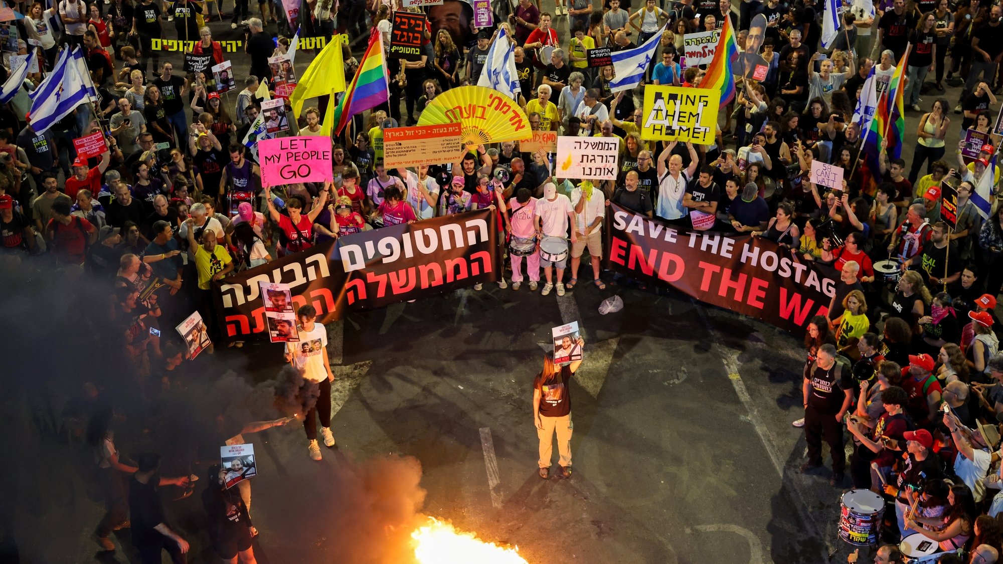 Multitud en una protesta con pancartas, algunos encendiendo fuego en el suelo. Energía intensa y decidida.