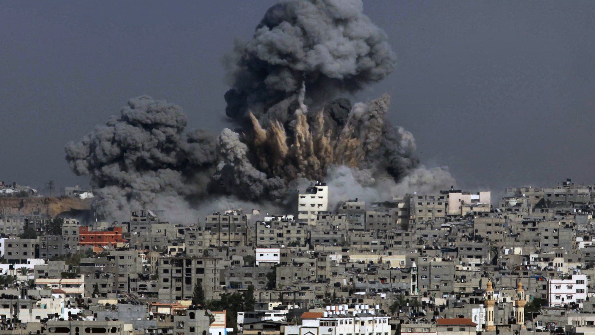 La imagen muestra una explosión en un área urbana, donde se elevan grandes nubes de humo y escombros hacia el cielo. Las estructuras de edificios en la parte inferior parecen estar dañadas, y el entorno refleja un ambiente de conflicto o desastre. La atmósfera es de caos, con el cielo grisáceo debido al humo de la explosión.