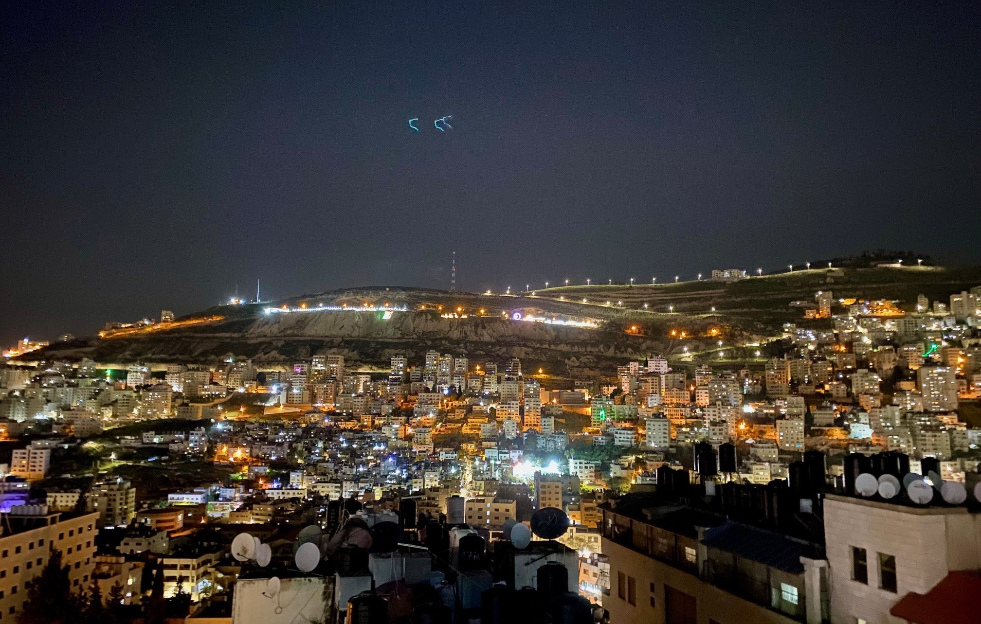 La imagen muestra una vista nocturna de la ciudad, iluminada por luces de diversos colores. Se pueden ver edificios y viviendas en la parte inferior, con antenas en los techos de algunas estructuras. En el fondo, hay colinas que también están iluminadas, y el cielo es oscuro, lo que resalta aún más las luces de la ciudad. Es un paisaje urbano que refleja la actividad y la vida nocturna del lugar.