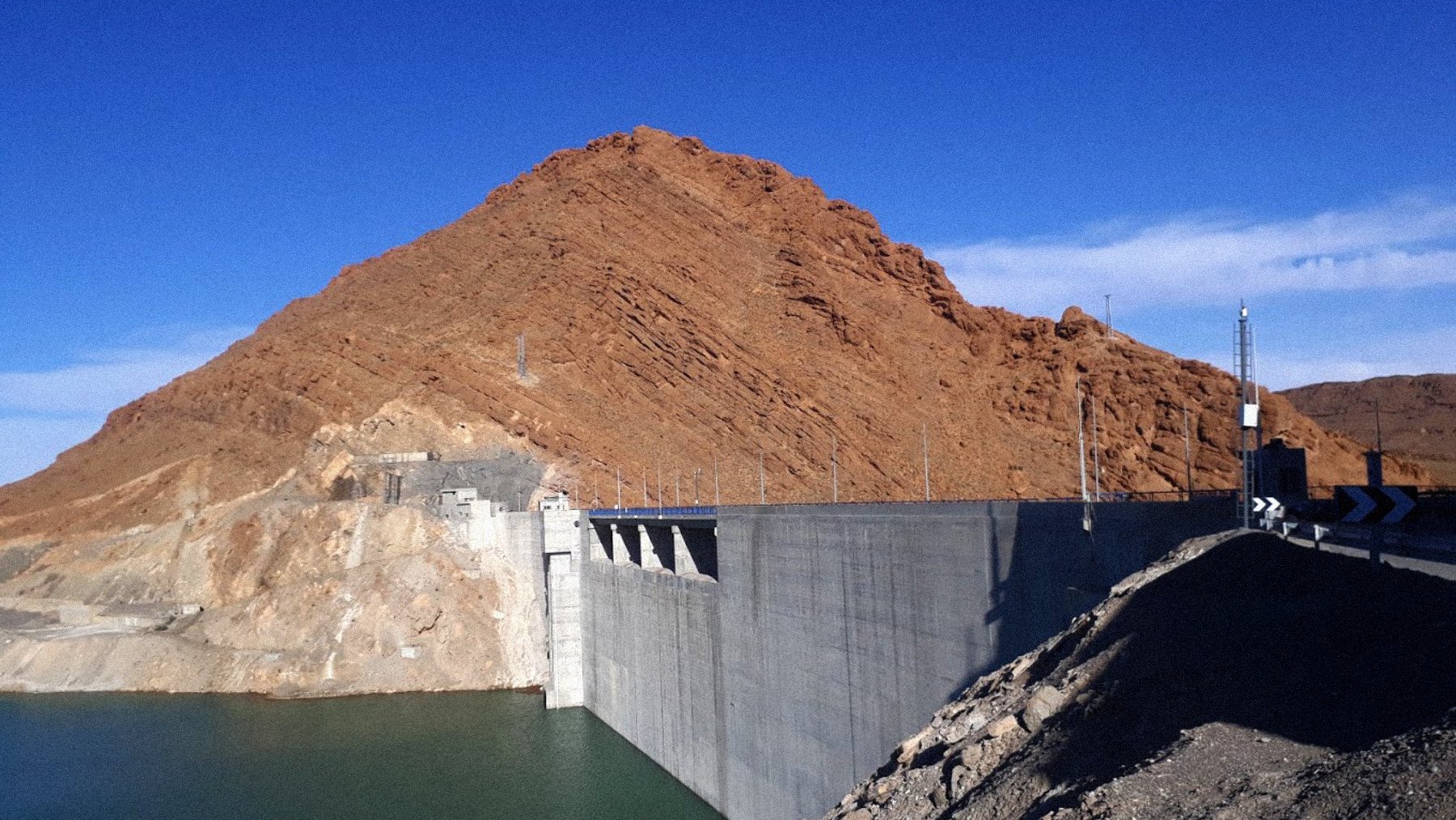 La imagen muestra un gran embalse o represa construida de concreto, situada a la orilla de un cuerpo de agua de color verde. Detrás de la represa se puede ver una montaña de tonalidades marrones y naranjas, con un cielo despejado de un azul brillante en el fondo. La estructura de la represa es sólida y parece estar bien mantenida, mientras que el terreno alrededor tiene un aspecto árido y rocoso.