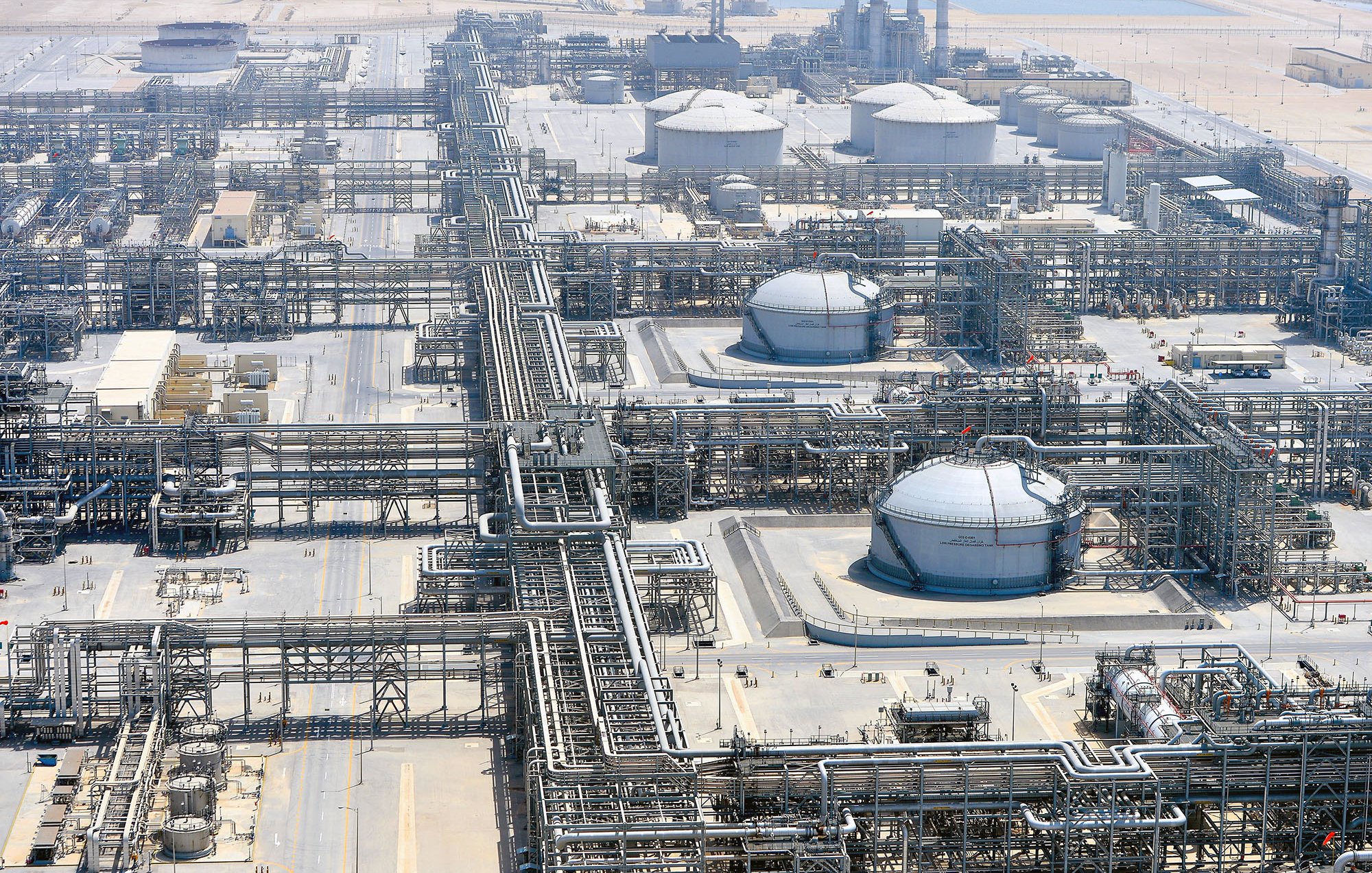 La imagen muestra una vista aérea de una planta industrial, probablemente relacionada con la producción o el refinado de petróleo. Se pueden observar grandes tanques de almacenamiento, tuberías extensas que se entrelazan por todo el área y estructuras de soporte. El entorno es desértico, con un paisaje árido al fondo, lo que sugiere que esta instalación podría estar ubicada en una región donde la extracción de recursos naturales es común. La organización y complejidad de las tuberías y tanques reflejan la sofisticación de la infraestructura industrial.