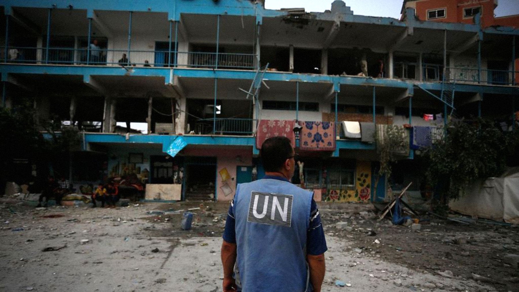 The image depicts a person wearing a blue vest labeled "UN," standing in front of a damaged building. The structure shows signs of destruction, with broken windows and debris scattered around the ground. There are individuals visible in the background, and the overall scene conveys a somber atmosphere, likely in a conflict or disaster-stricken area.